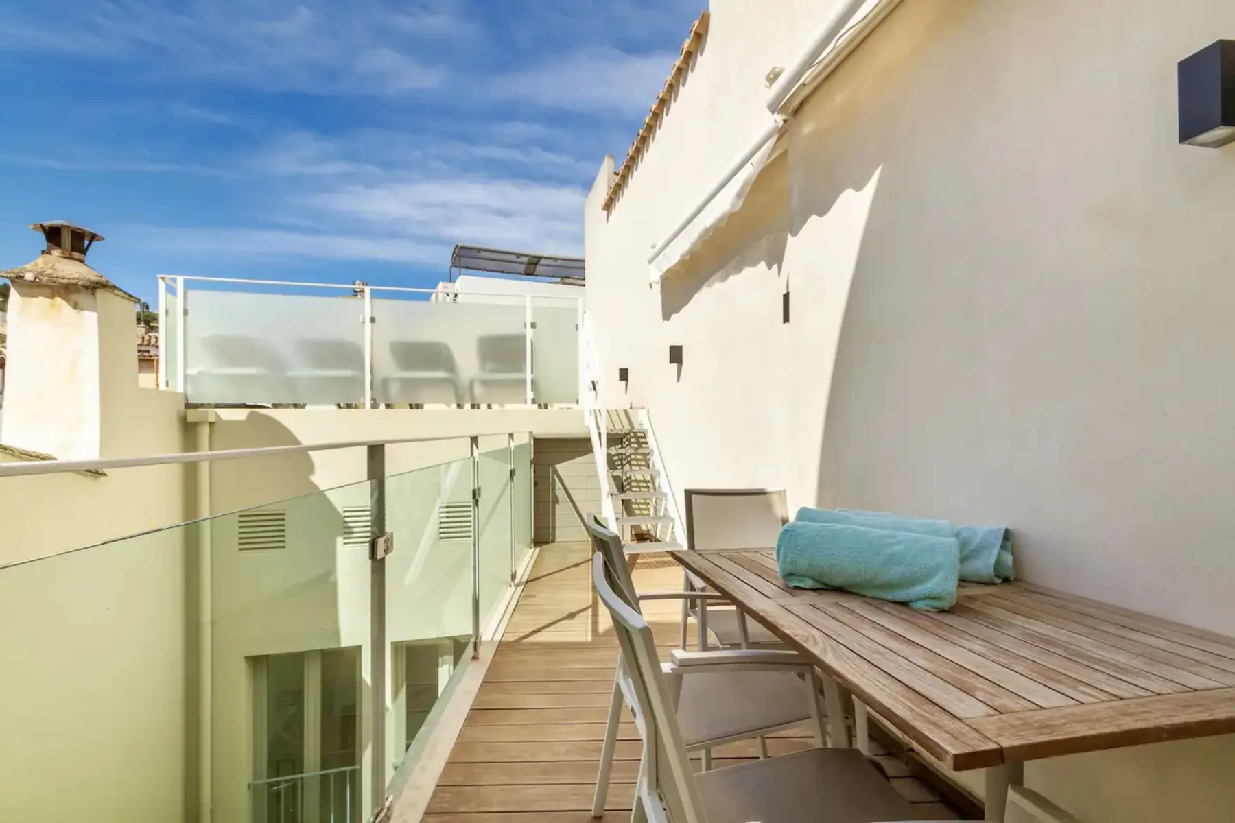 Una terraza soleada tiene una mesa de comedor de madera y sillas junto a barandillas de vidrio.