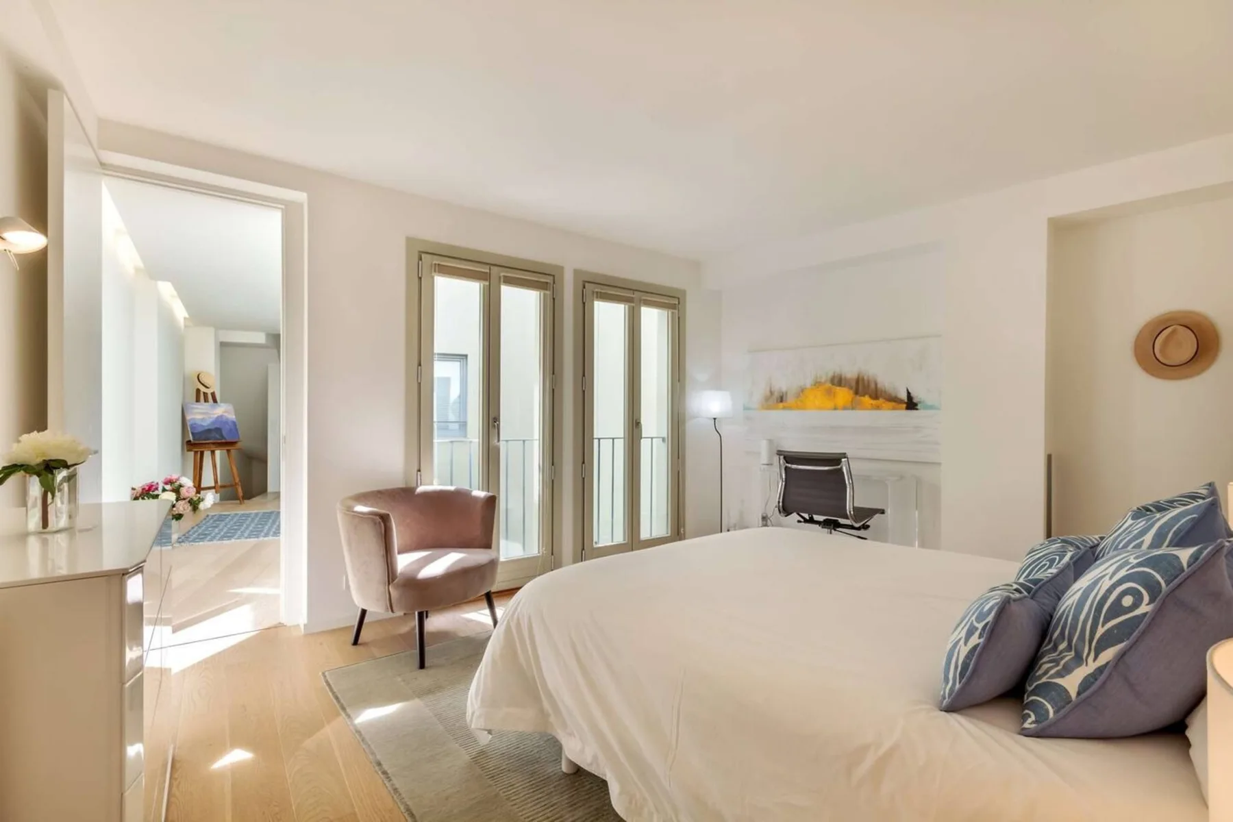 Un dormitorio luminoso con una cama grande, un sillón y ventanas altas.