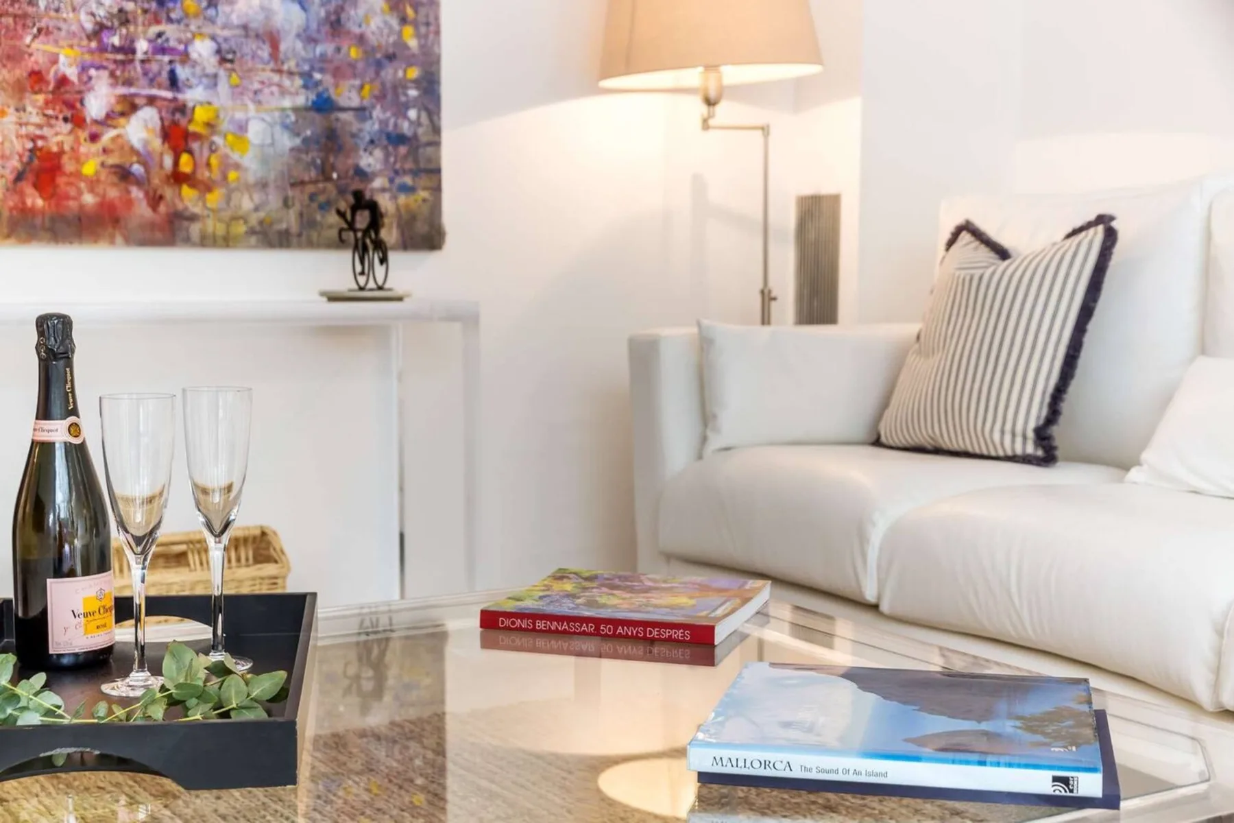 Una sala de estar luminosa con asientos blancos, una mesa de centro de vidrio y arte colorido.