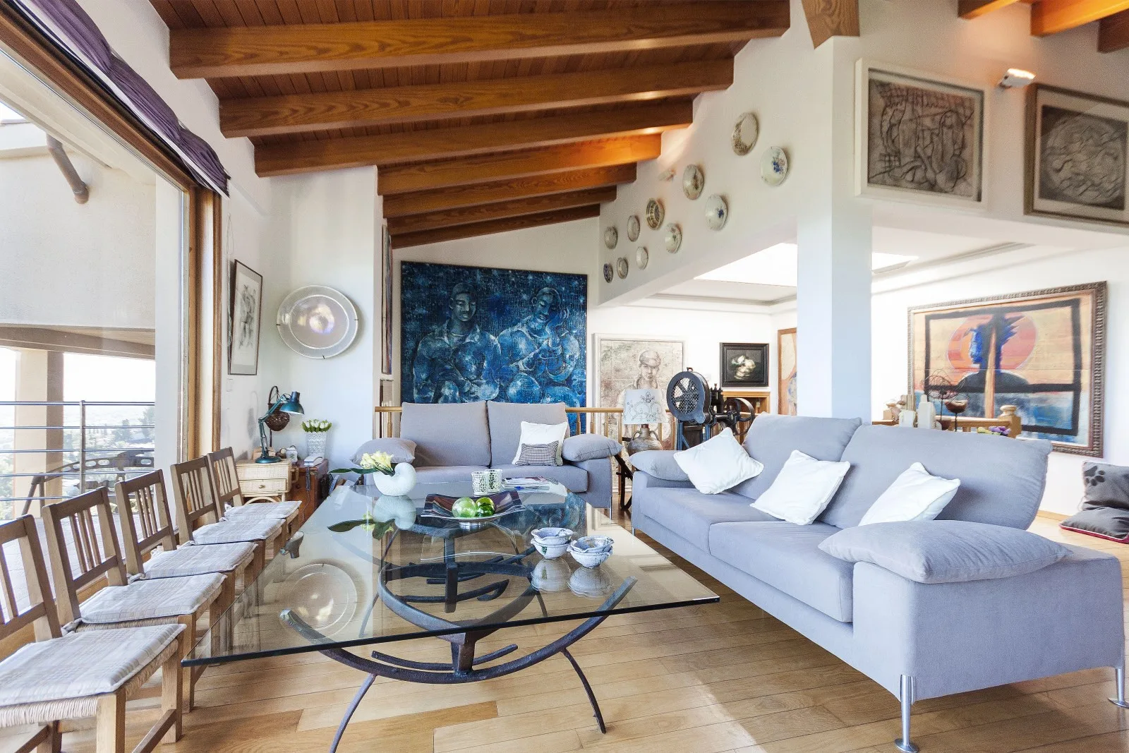 A bright living room with gray seating, a glass coffee table, and exposed wooden ceiling beams.