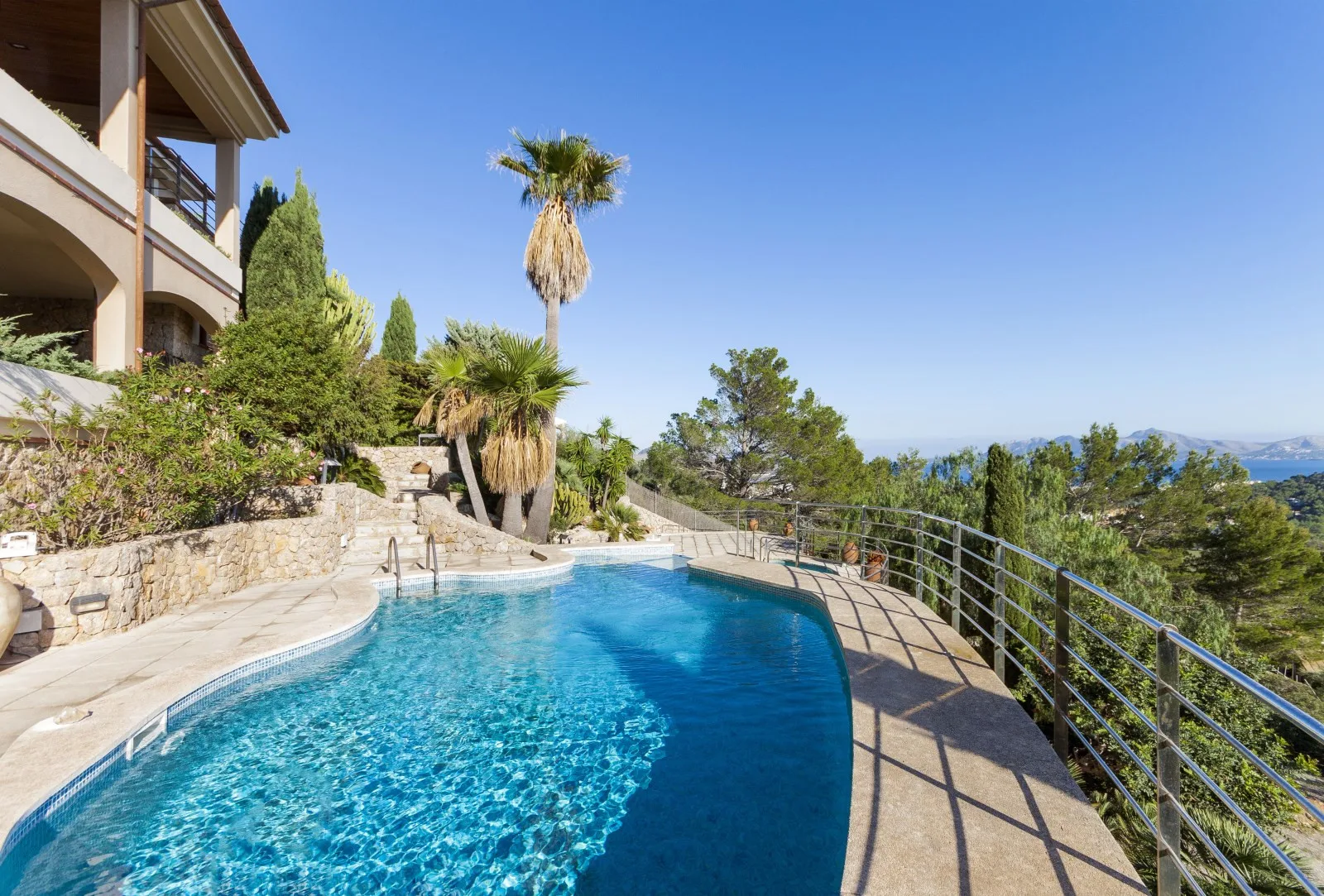 A curved outdoor swimming pool overlooks a tree-covered landscape beneath a clear blue sky.