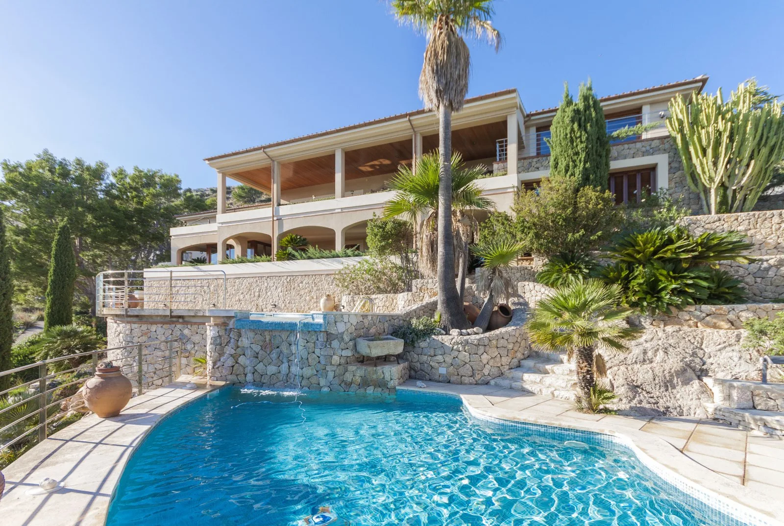 A swimming pool sits below a large house on a sunny, landscaped hillside.