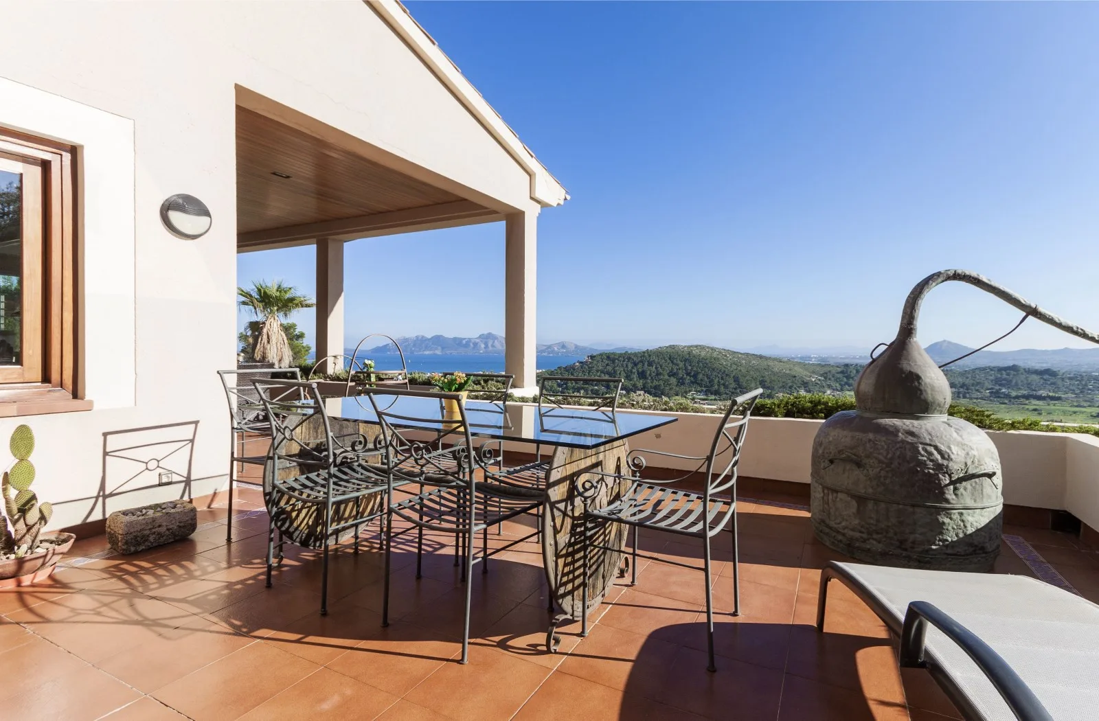 A sunny outdoor terrace has a dining table and chairs overlooking a coastal landscape.