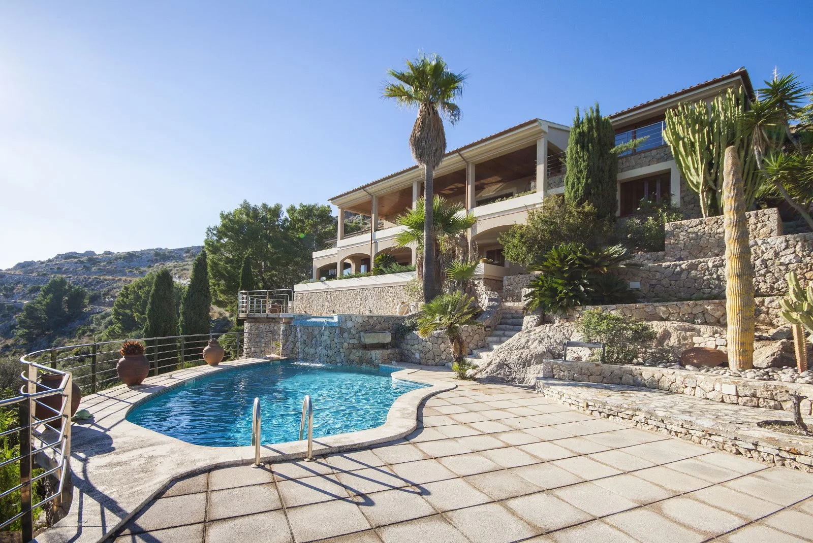 A hillside villa overlooks a swimming pool and paved terrace.
