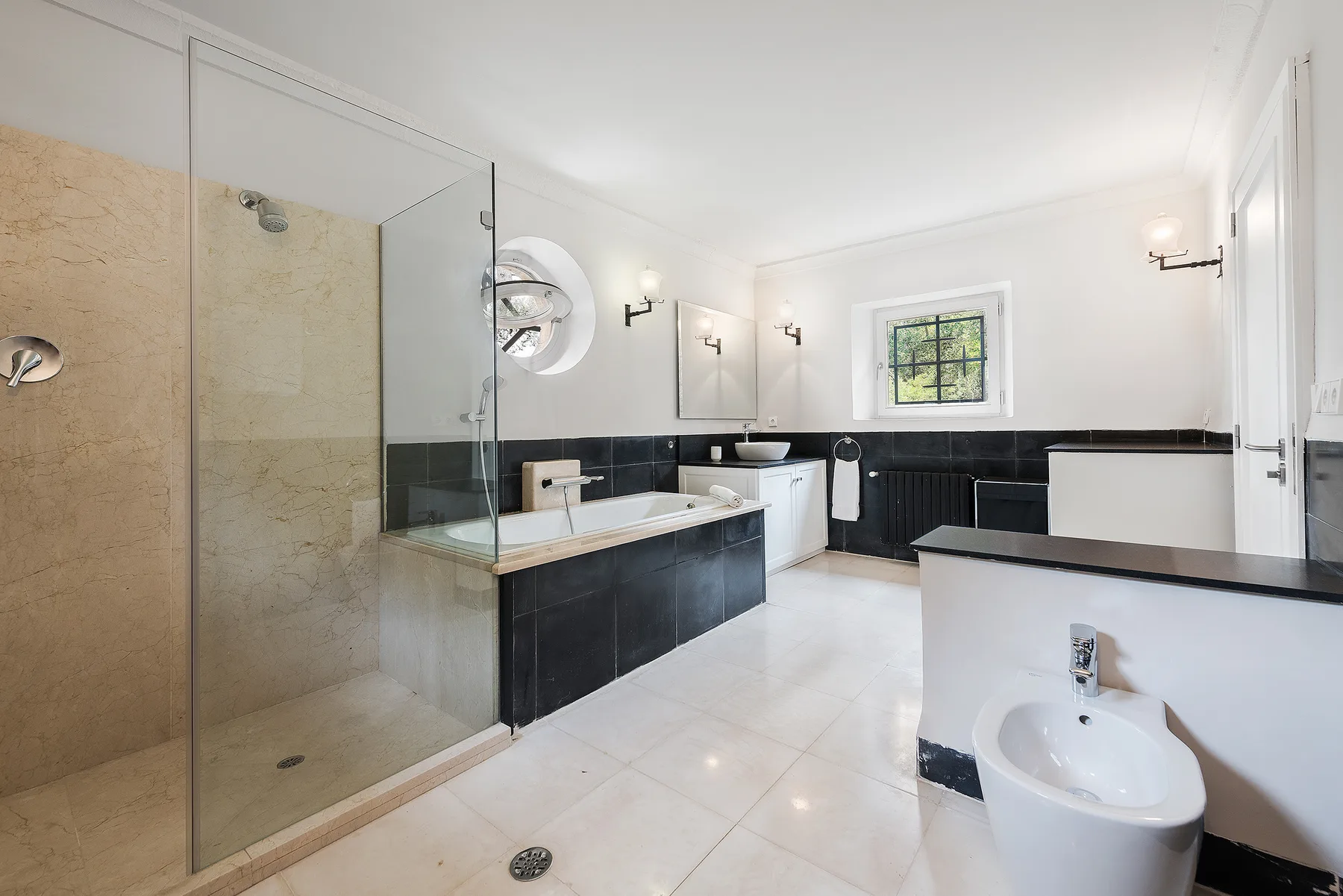 A bright bathroom with a glass shower, dark tiled walls, and a window.