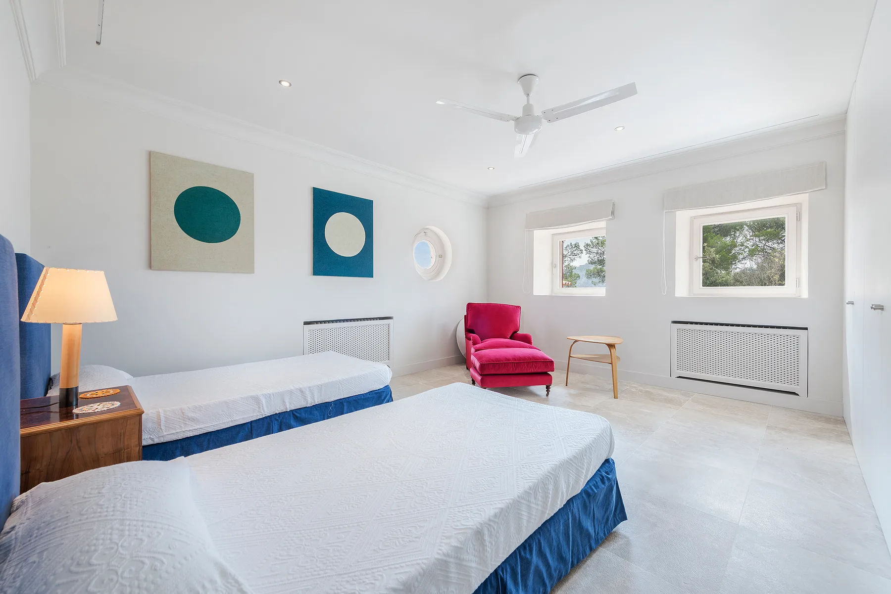 A bright bedroom with two beds, a red armchair, and windows.