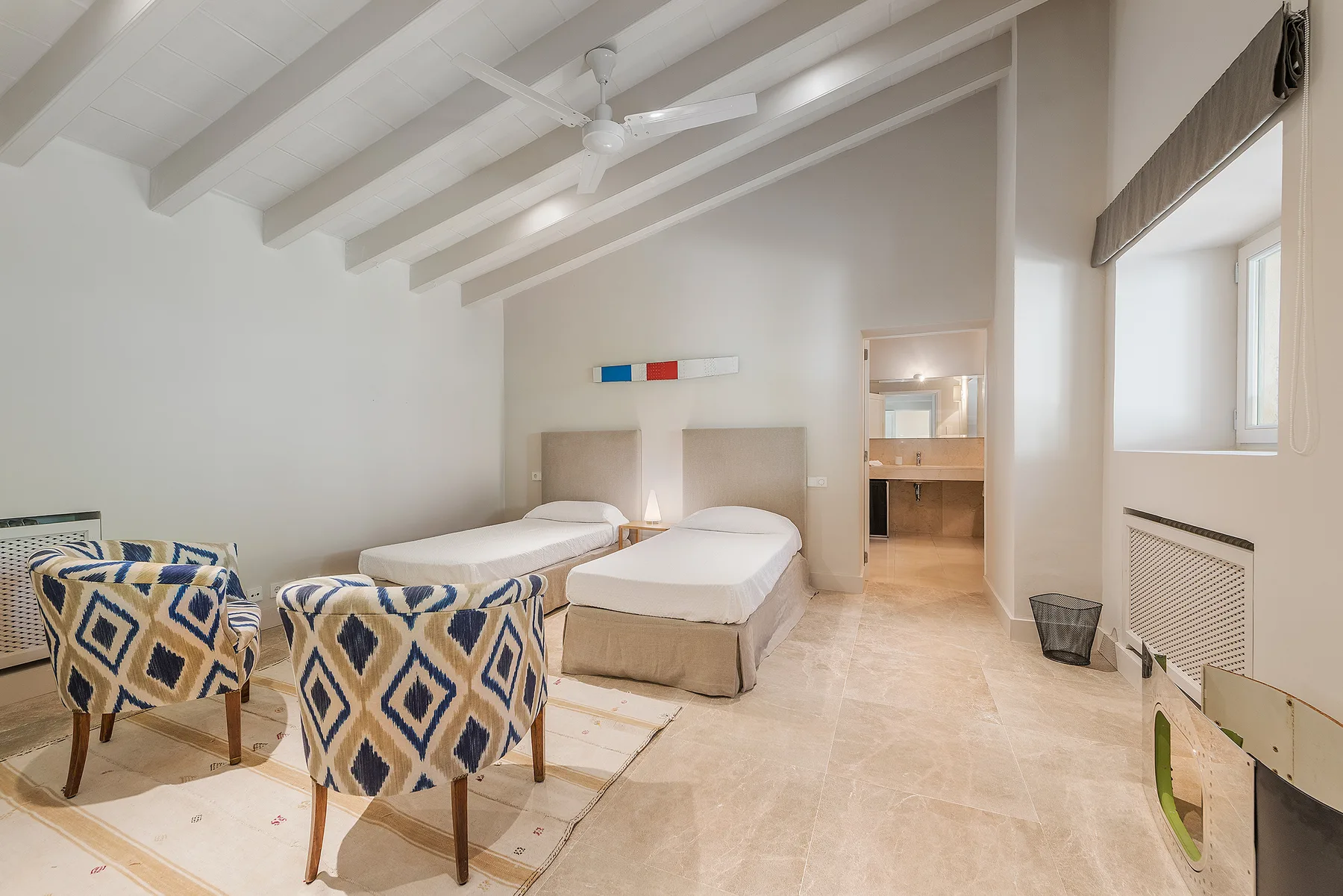 A bright bedroom with two beds and patterned seating beneath a vaulted ceiling.