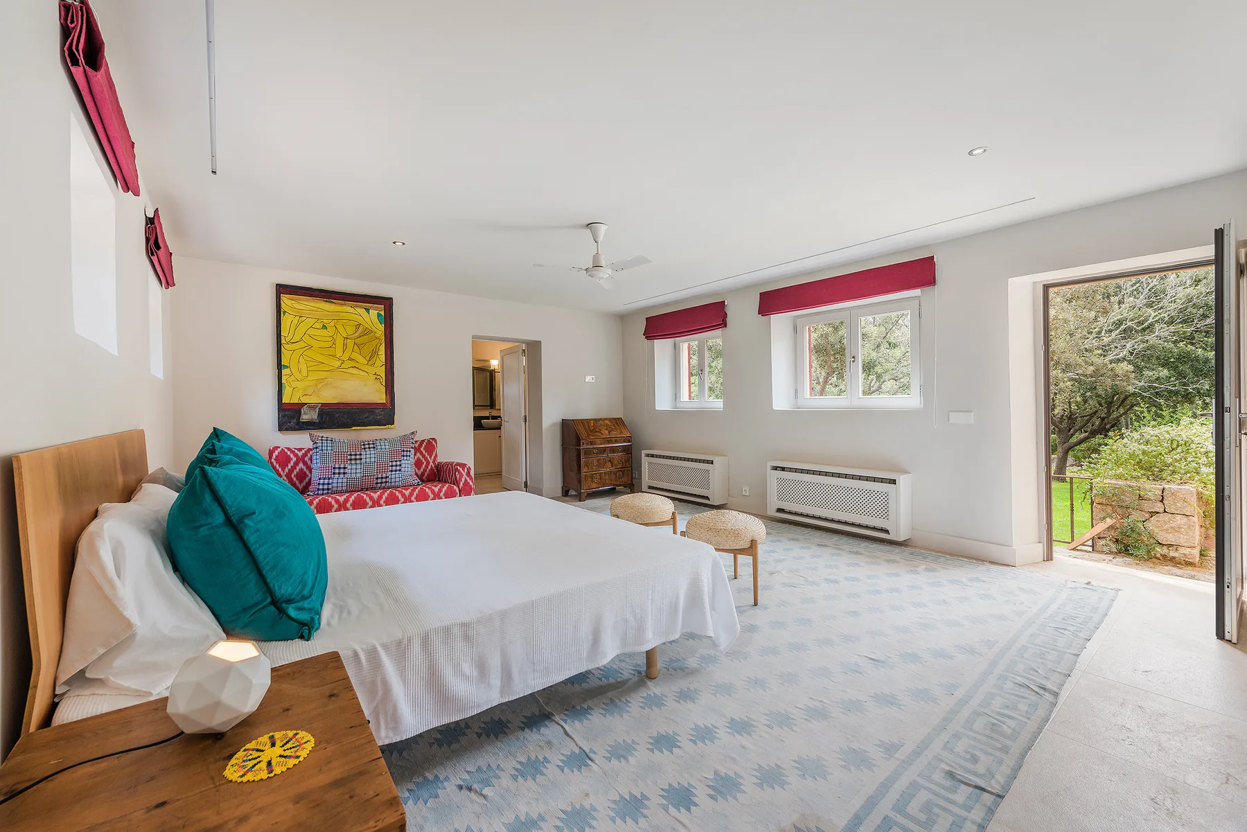 A bedroom with a large bed, colorful cushions, and a glass door opening to greenery.