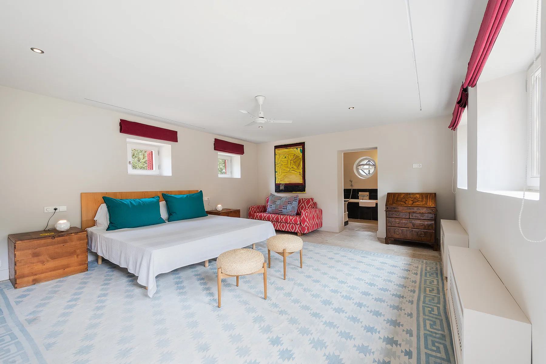 A bright bedroom with a bed, patterned floor covering, and large windows.
