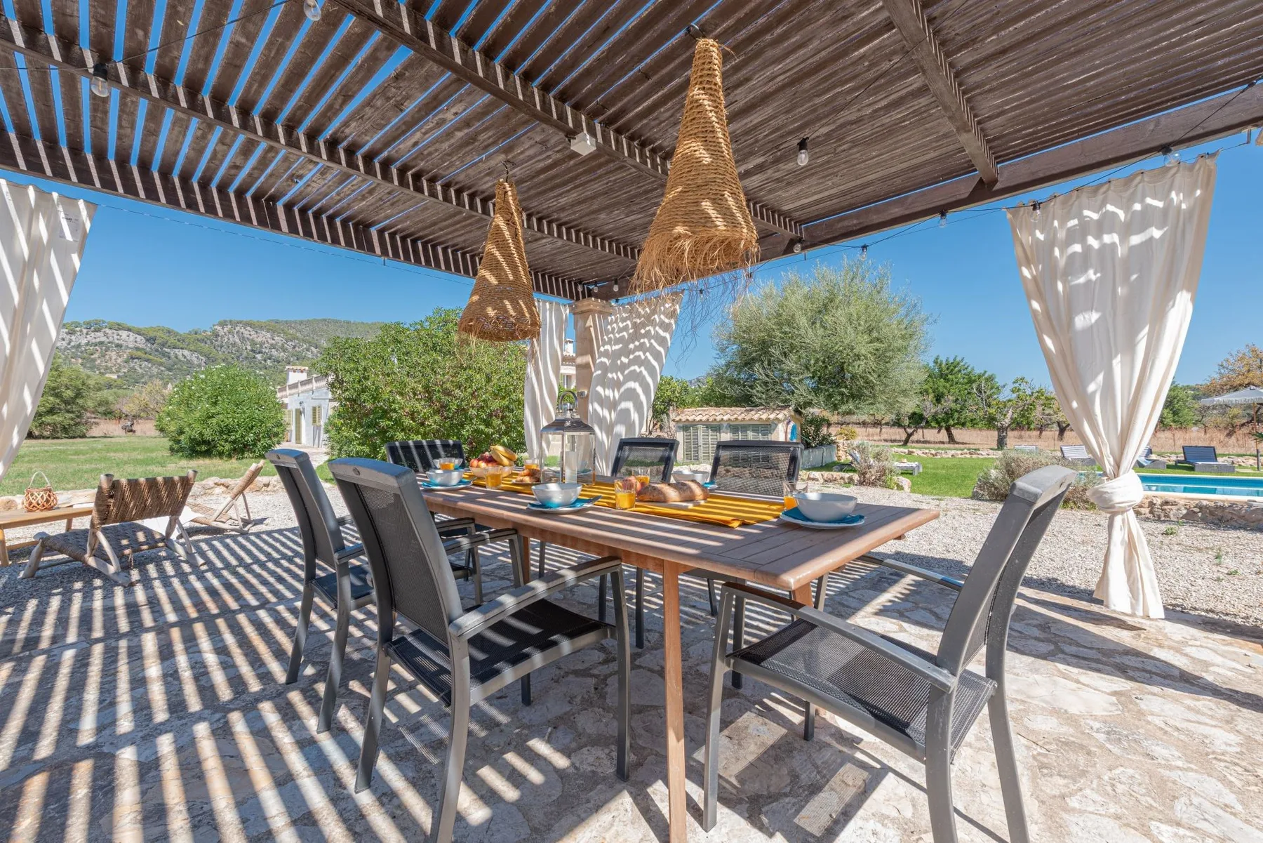 Una zona de comedor exterior a la sombra, con la mesa puesta bajo una pérgola de madera.