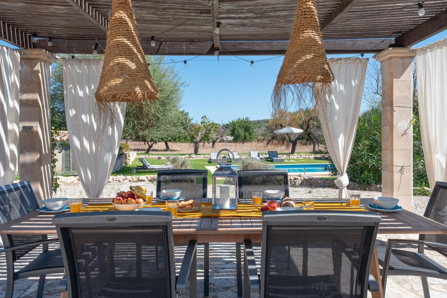 Un comedor al aire libre bajo una pérgola da a una piscina y un jardín.