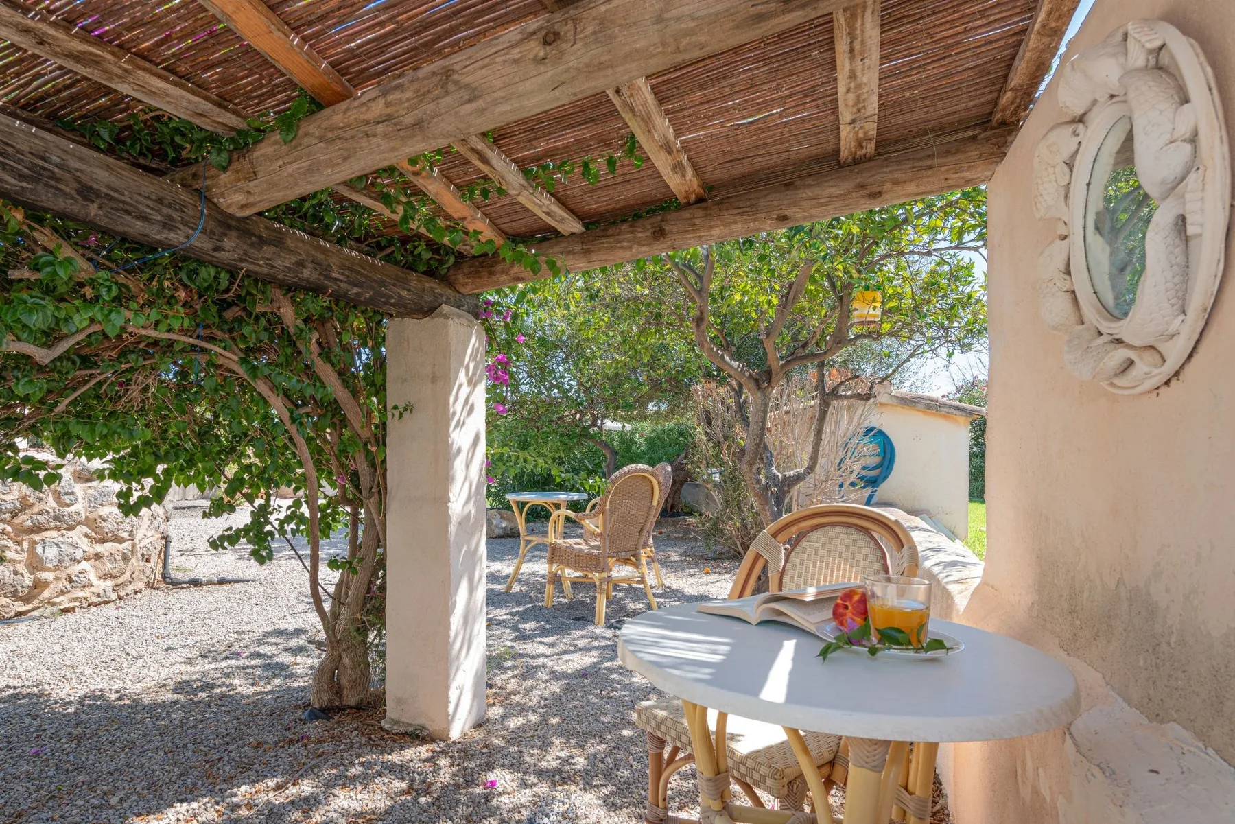 Zona de comedor exterior a la sombra, con mesa y asientos de mimbre bajo una pérgola cubierta de enredaderas.