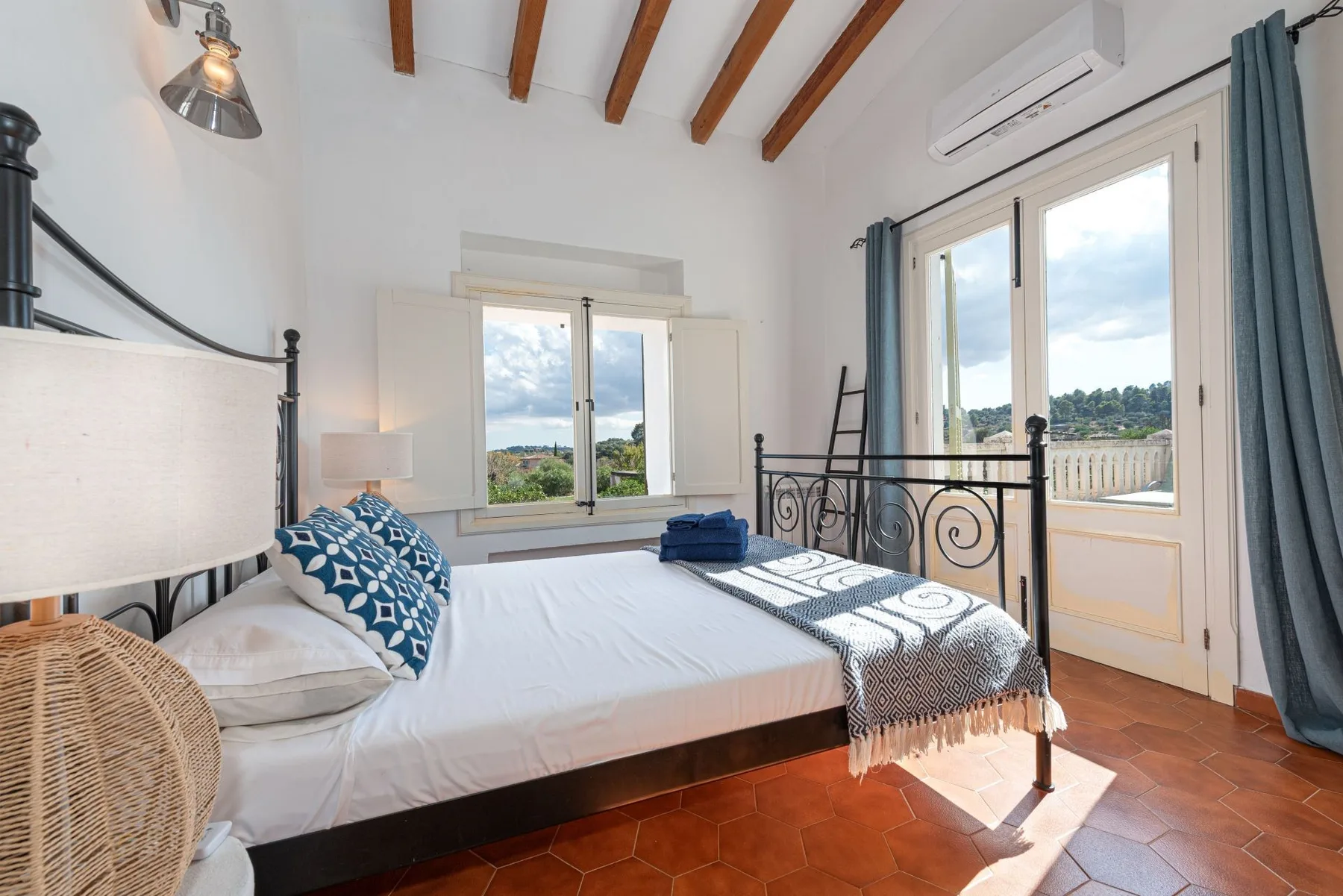 Un dormitorio luminoso con una cama junto a ventanas y una puerta acristalada que da a un balcón.