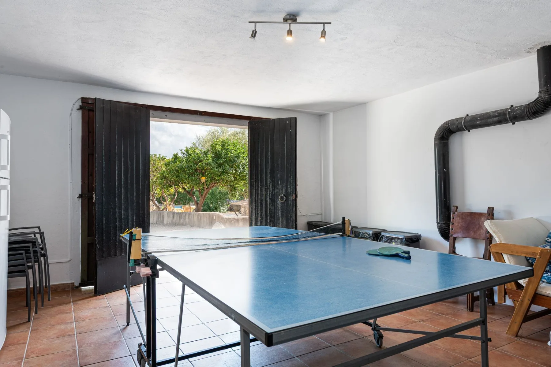 Una mesa de ping-pong ocupa una sala luminosa con puertas abiertas hacia una zona exterior arbolada.