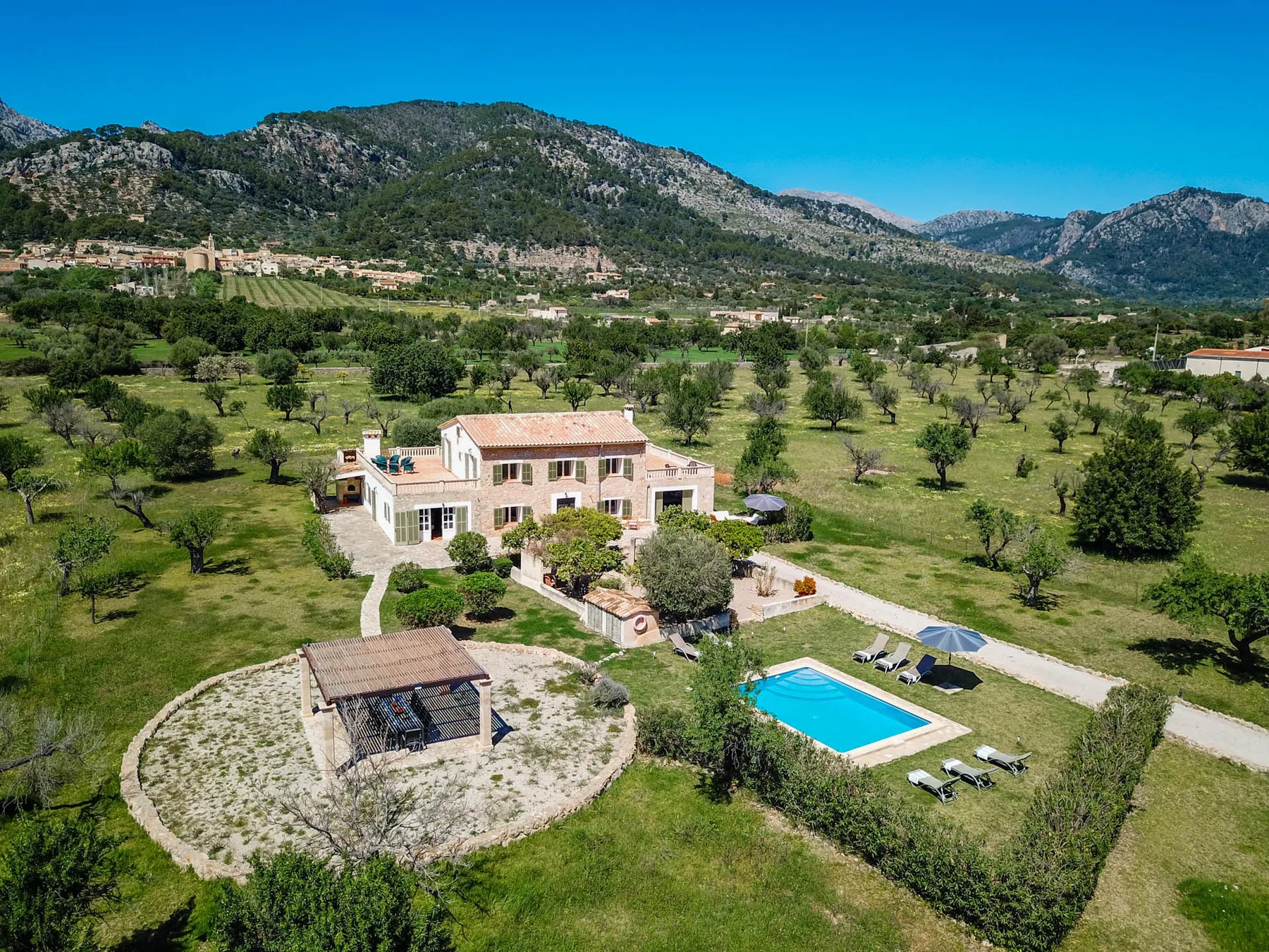 Una casa rural y una piscina se encuentran entre campos verdes, bajo una cadena montañosa.