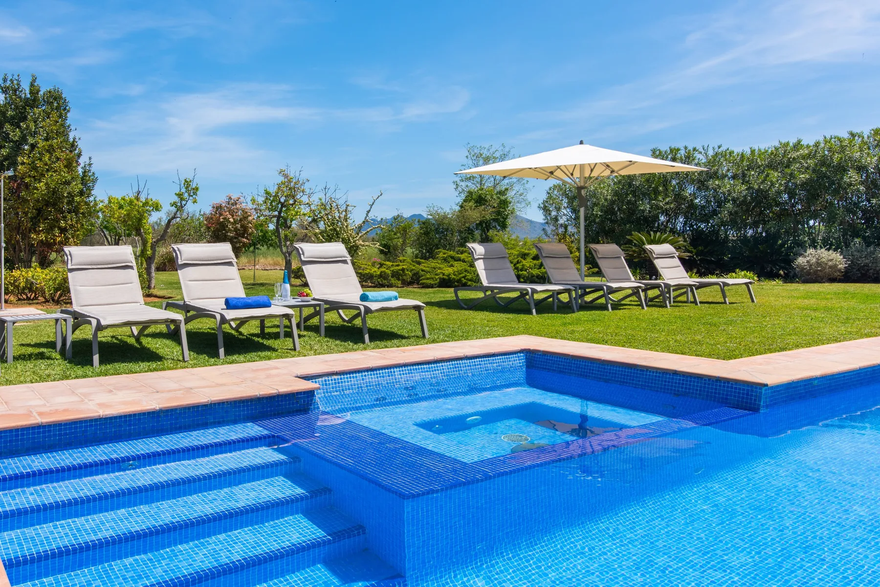 An outdoor swimming pool with steps, surrounded by loungers and greenery.
