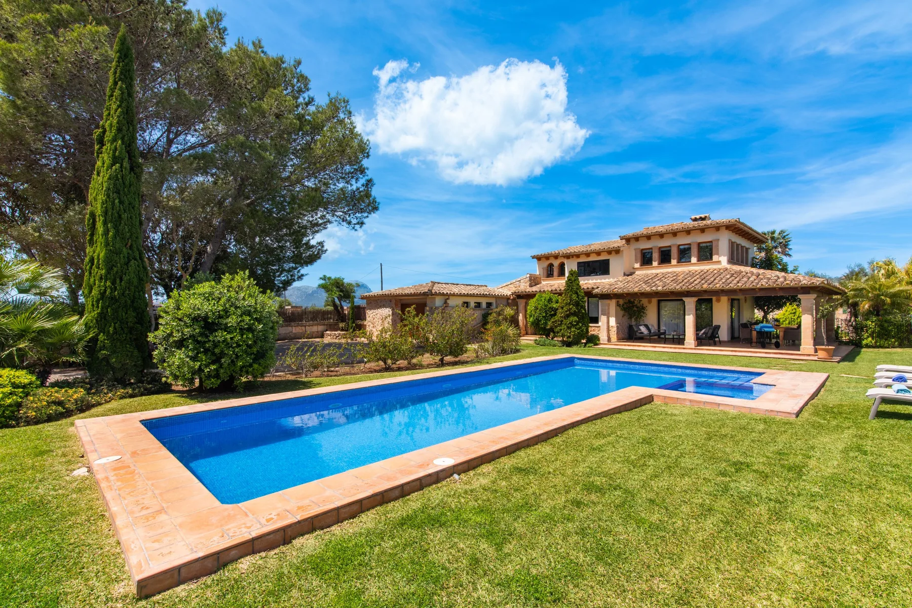 A swimming pool sits beside a lawn and a large house under a blue sky.