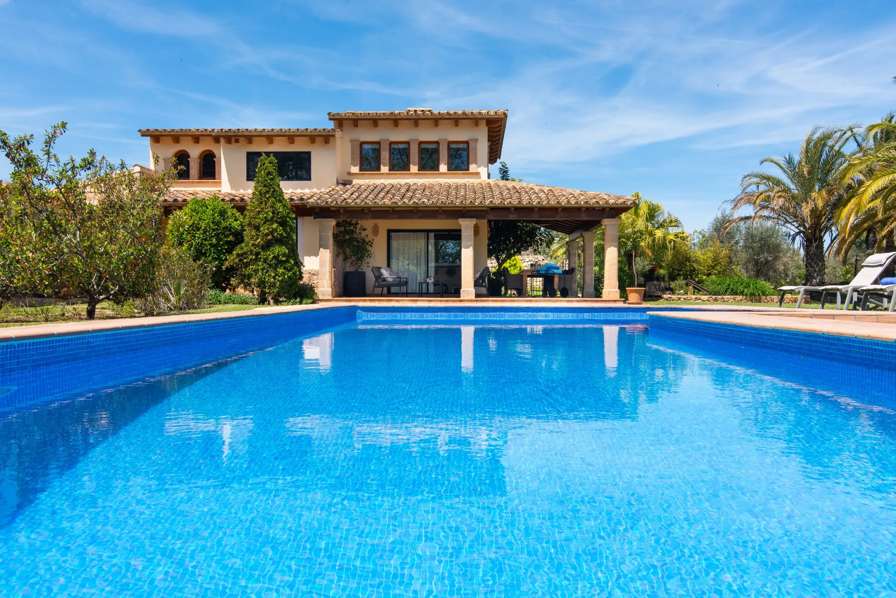 A swimming pool in front of a house, with patio seating and trees nearby.