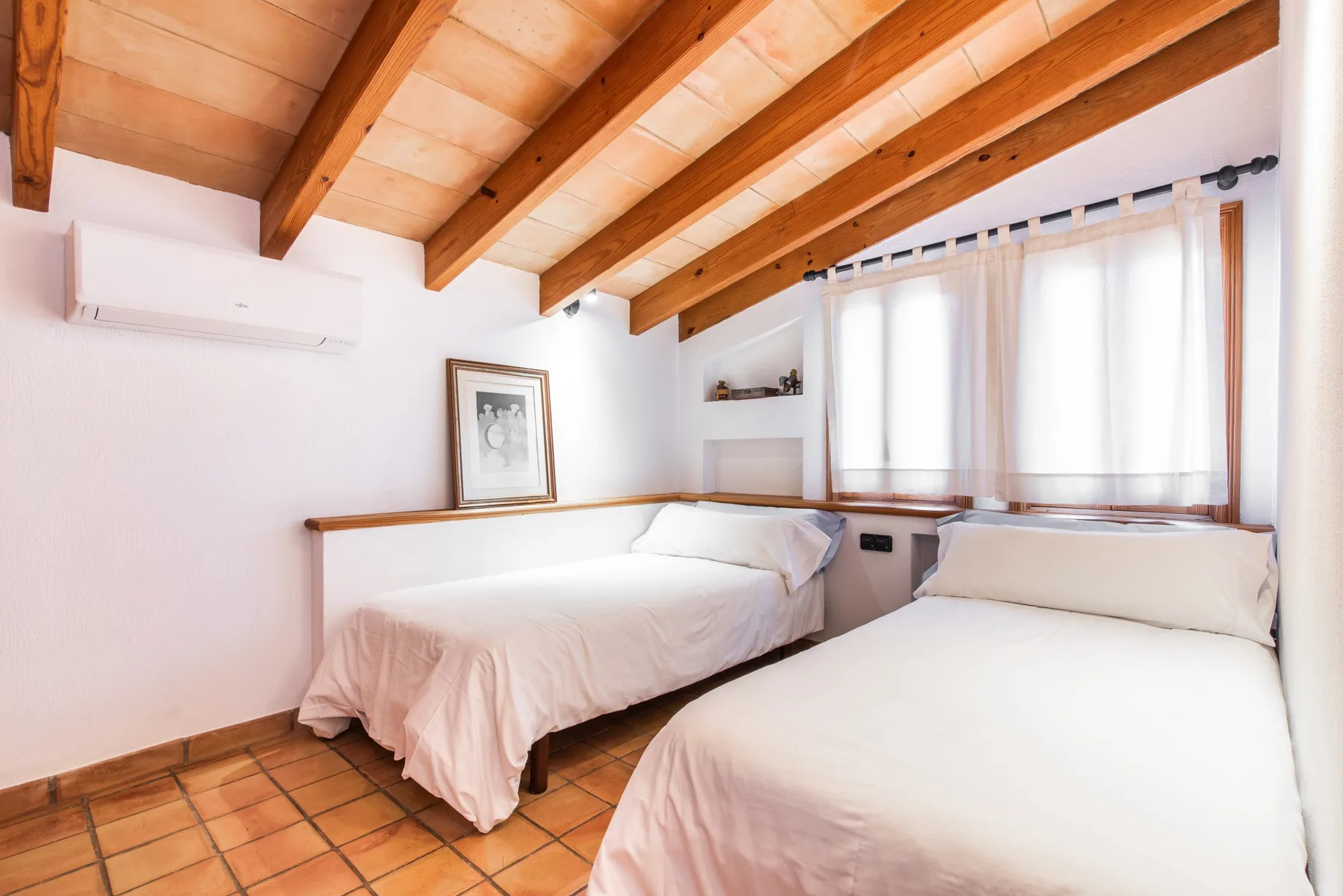 A bright bedroom with two beds beneath exposed wooden ceiling beams.