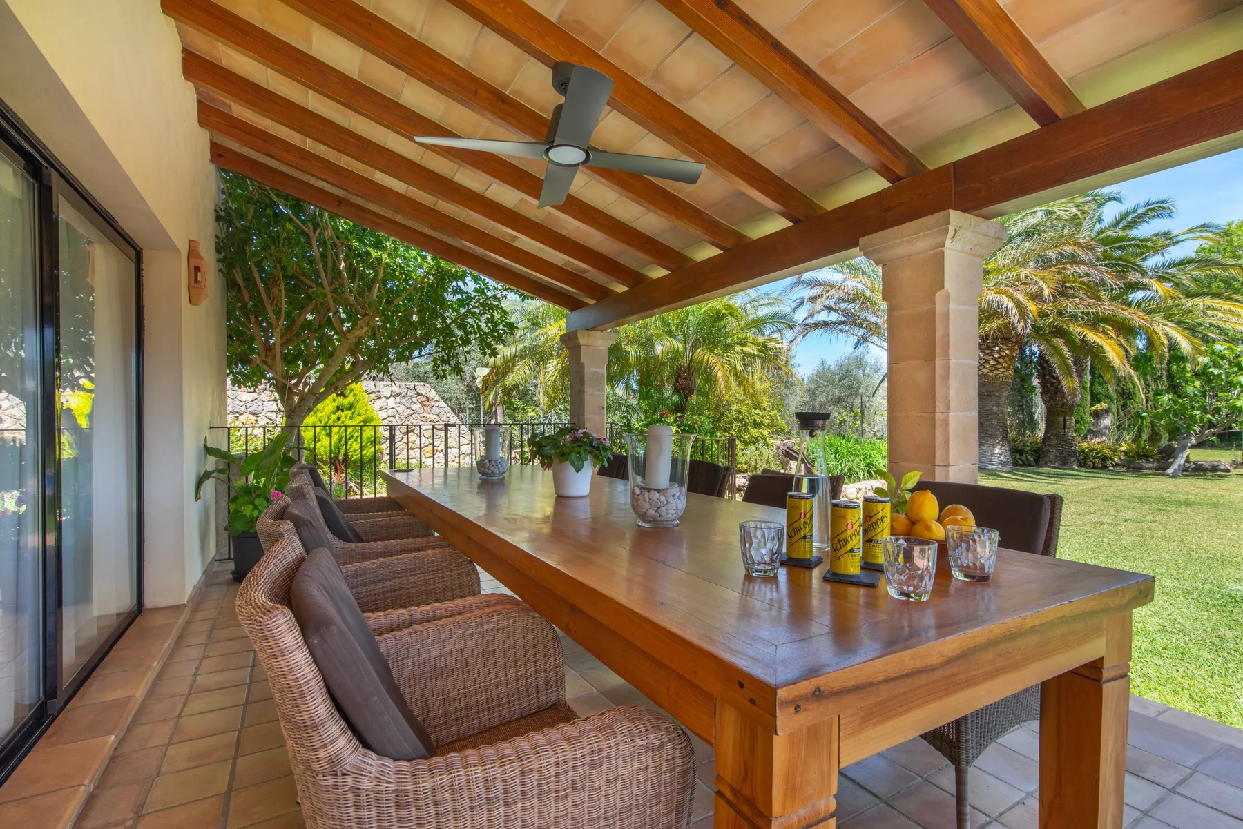 A covered outdoor dining area has a long wooden table and garden views.