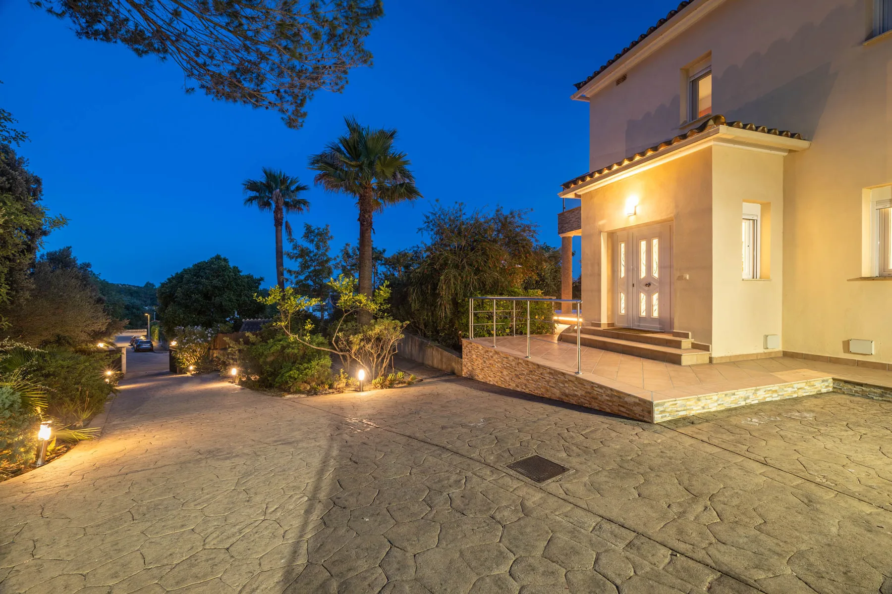 An illuminated house stands beside a paved courtyard and landscaped garden at dusk.