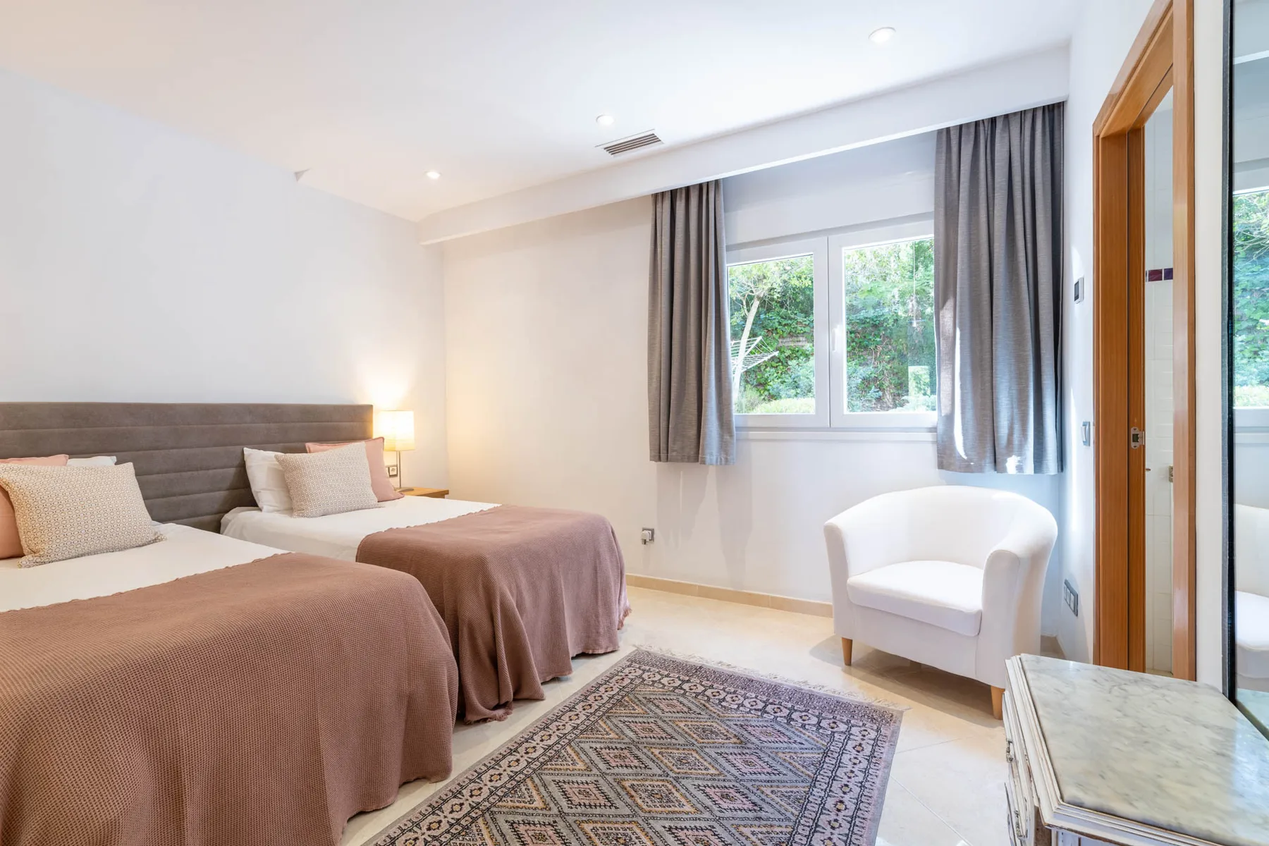 A bright bedroom with two beds, a window, and a white armchair.