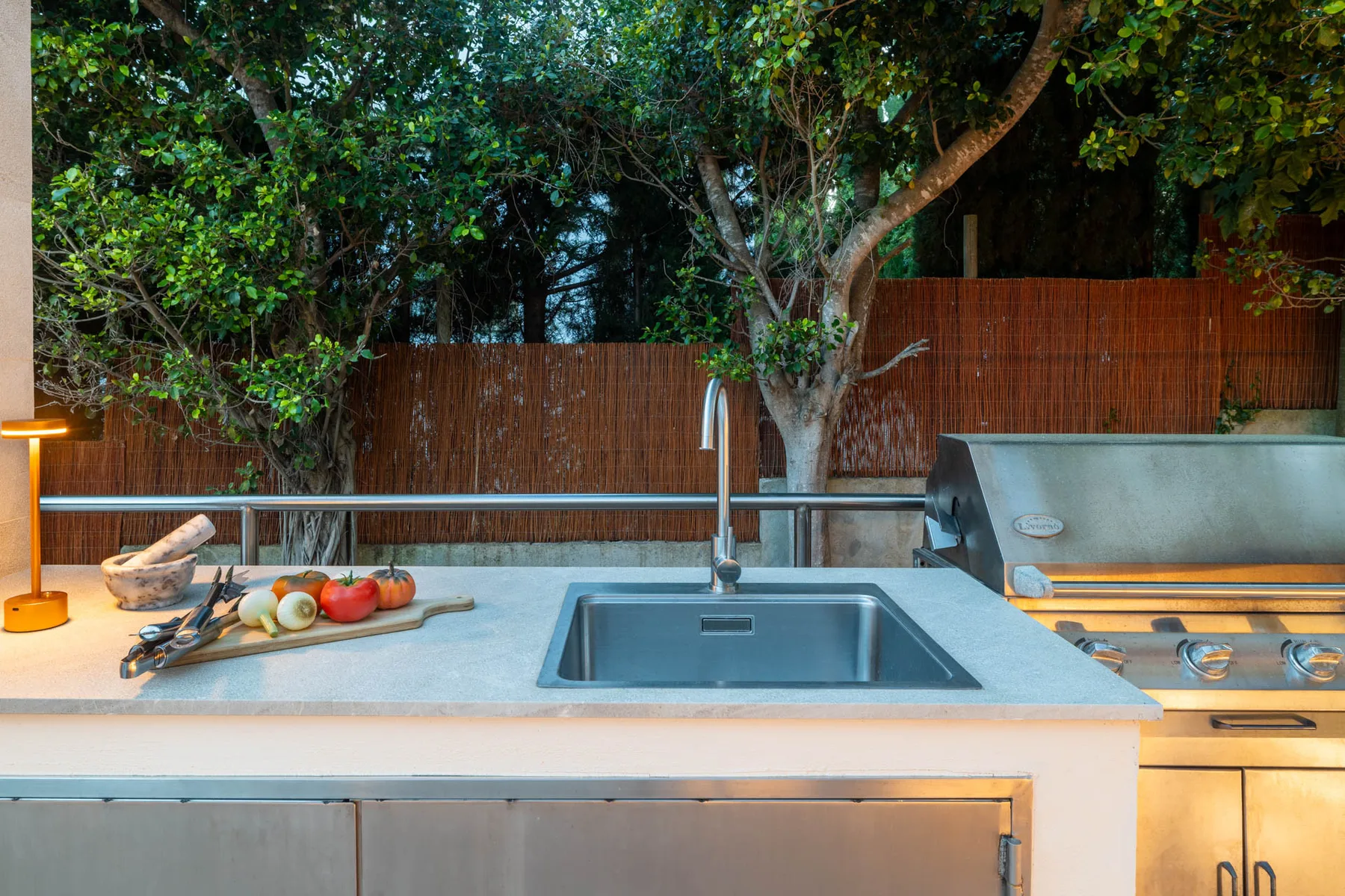 An outdoor kitchen counter with a sink and built-in grill beneath leafy trees.