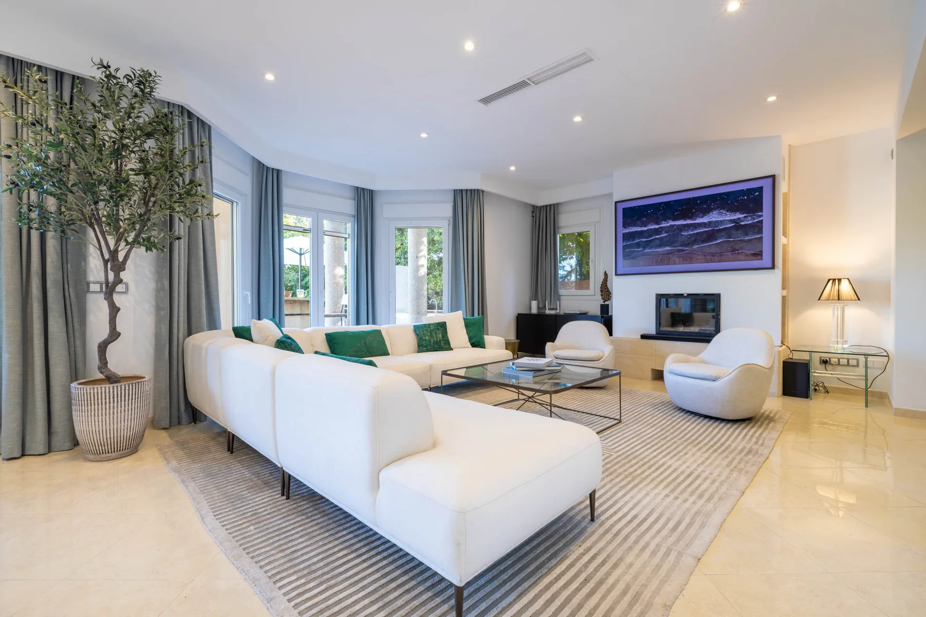 A bright living room with white seating, tall windows, and a wall-mounted television.