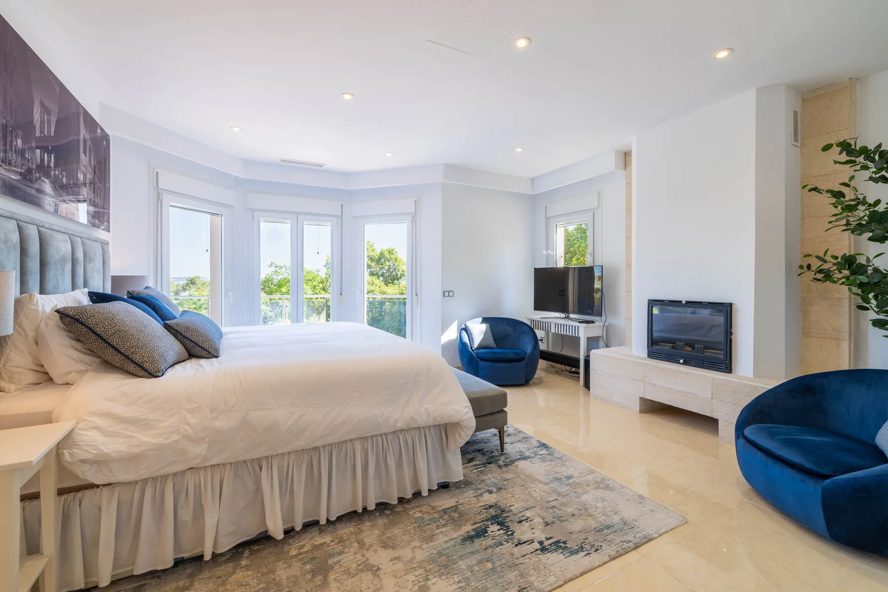 A bright bedroom with a large bed, blue seating, and windows overlooking greenery.