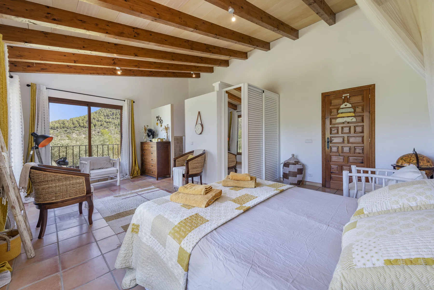 Un dormitorio luminoso con camas, vigas expuestas en el techo y un balcón con vistas a los árboles.