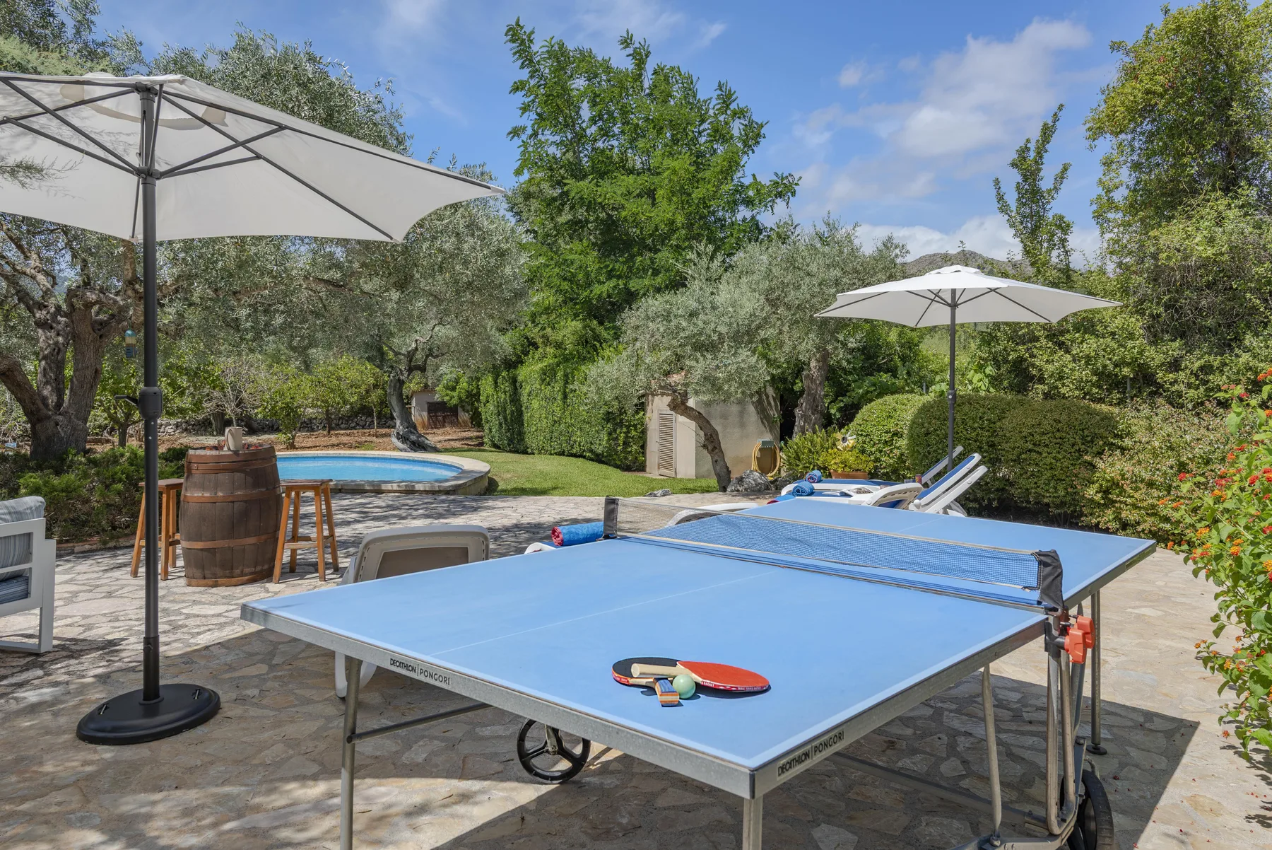 Una mesa de ping-pong está en un patio de piedra junto a una piscina pequeña y un jardín.