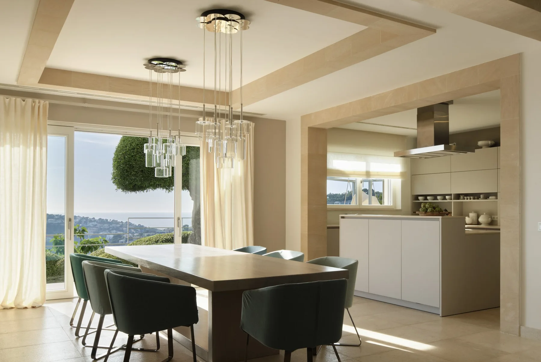 A dining area opens onto a kitchen, with a table and chairs beside large windows.