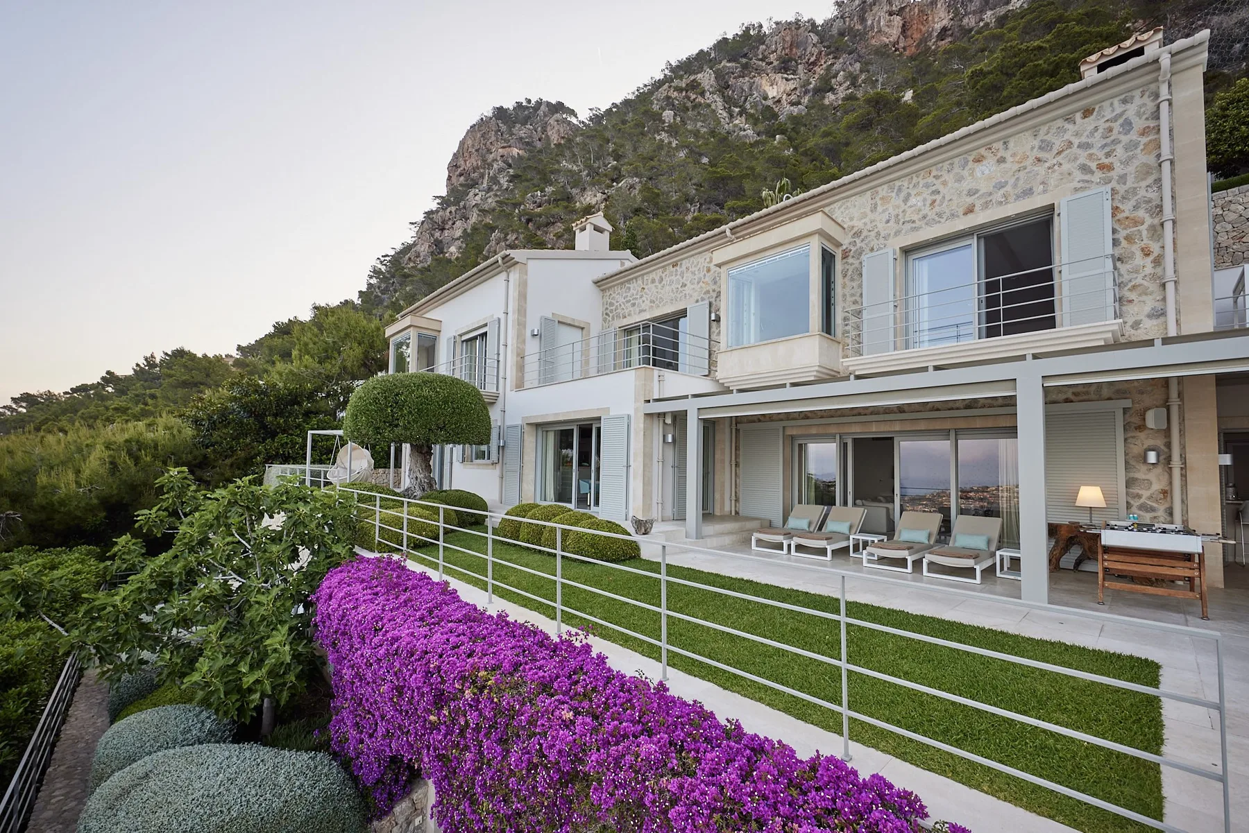 A modern stone-and-glass house overlooks a landscaped garden with purple flowers.