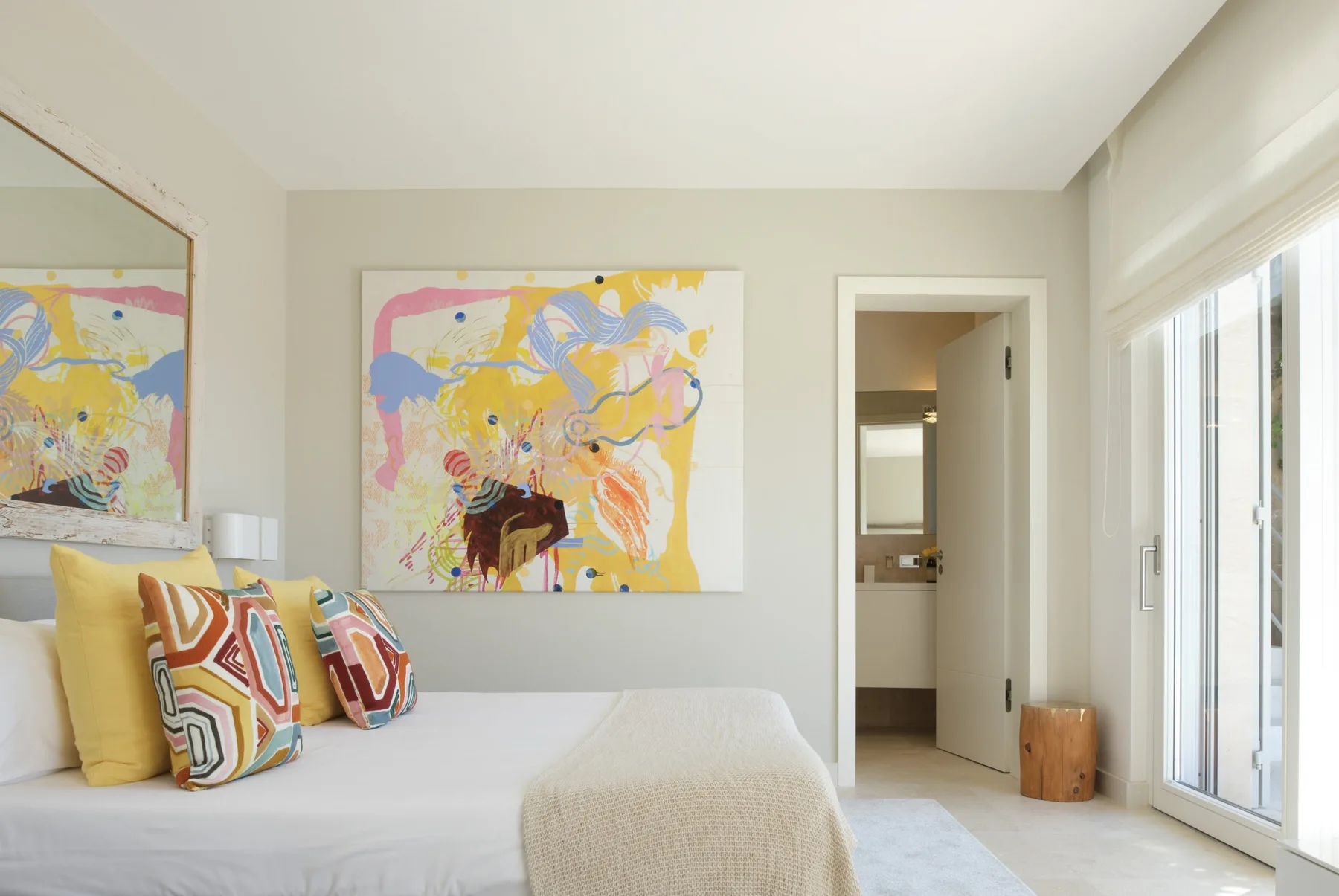 A bright bedroom with a bed, colorful artwork, and large windows.