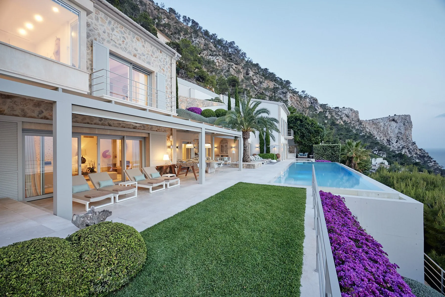 A modern hillside home opens onto a terrace, lawn, and long swimming pool.