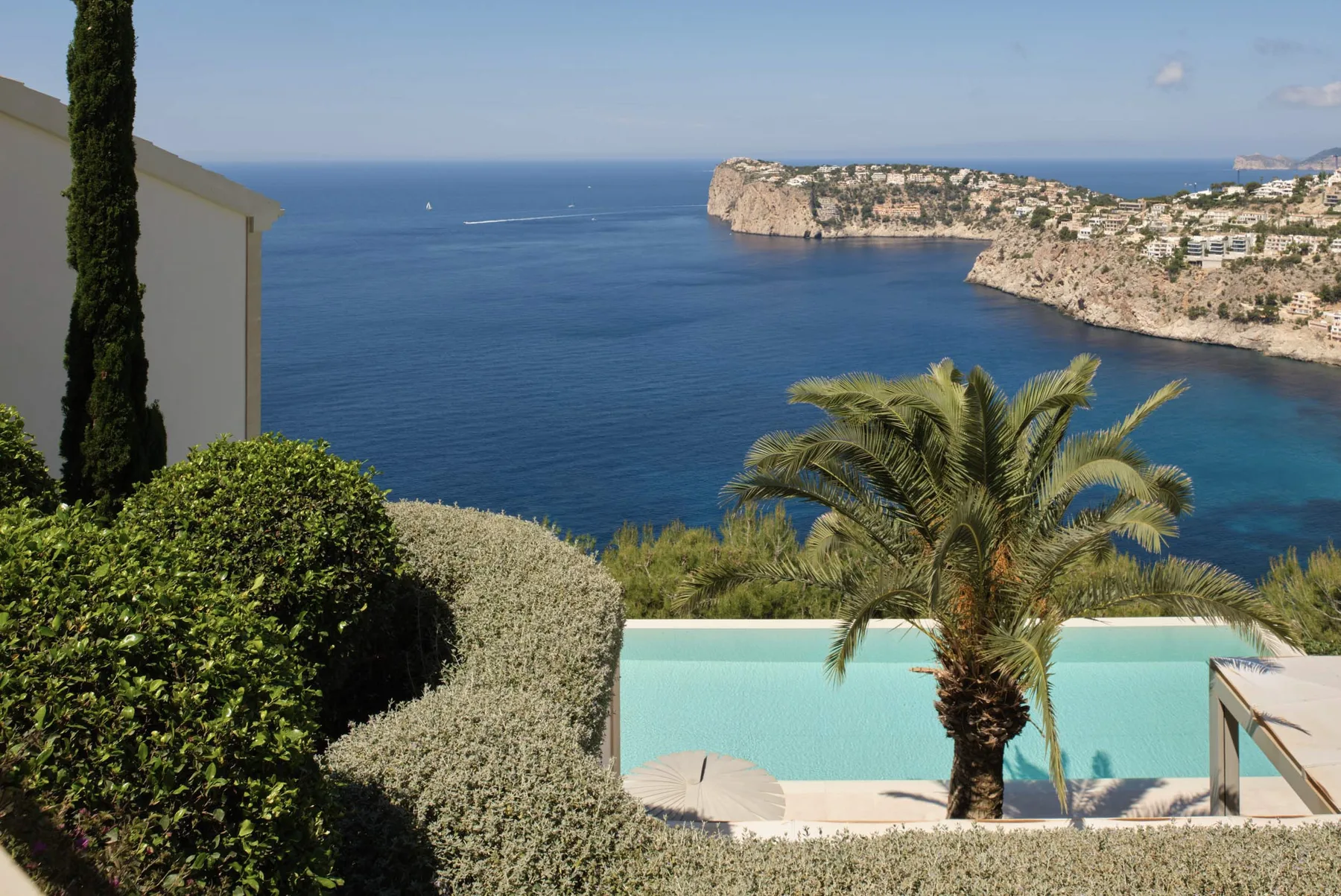 A swimming pool overlooks a blue bay, with palm trees and coastal cliffs.
