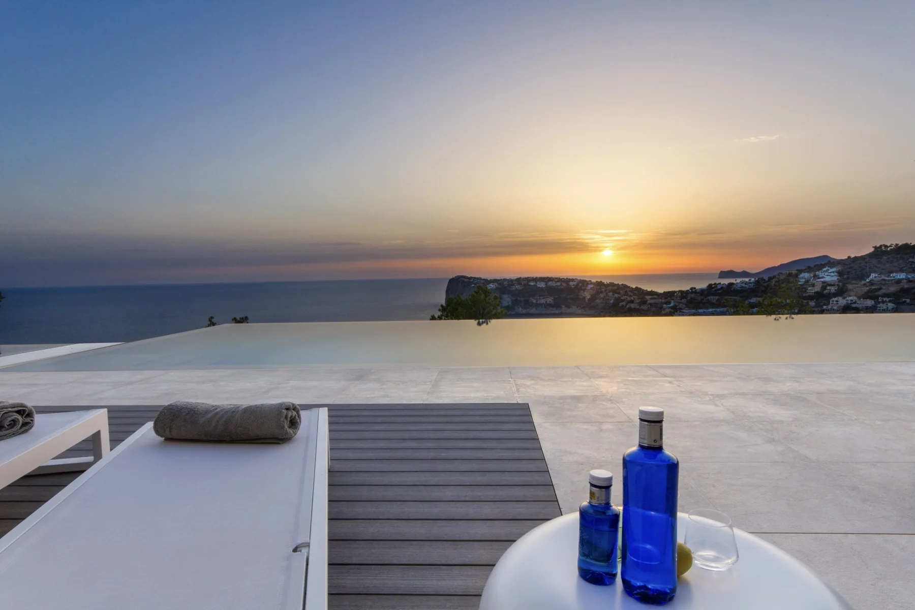 A rooftop terrace overlooks the sea at sunset, with bottles on a table.