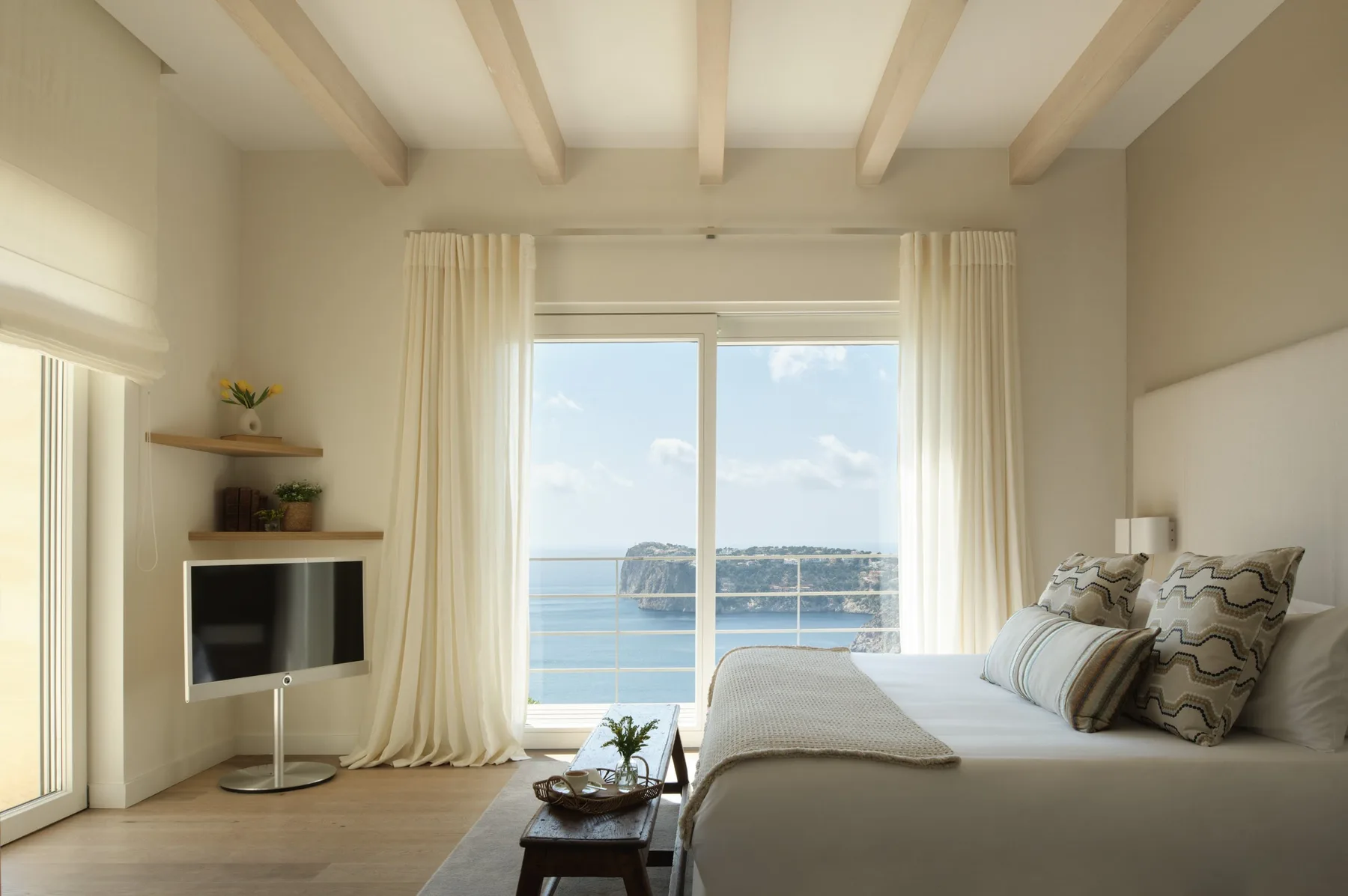 A bright bedroom with a bed beside a large window overlooking the sea.
