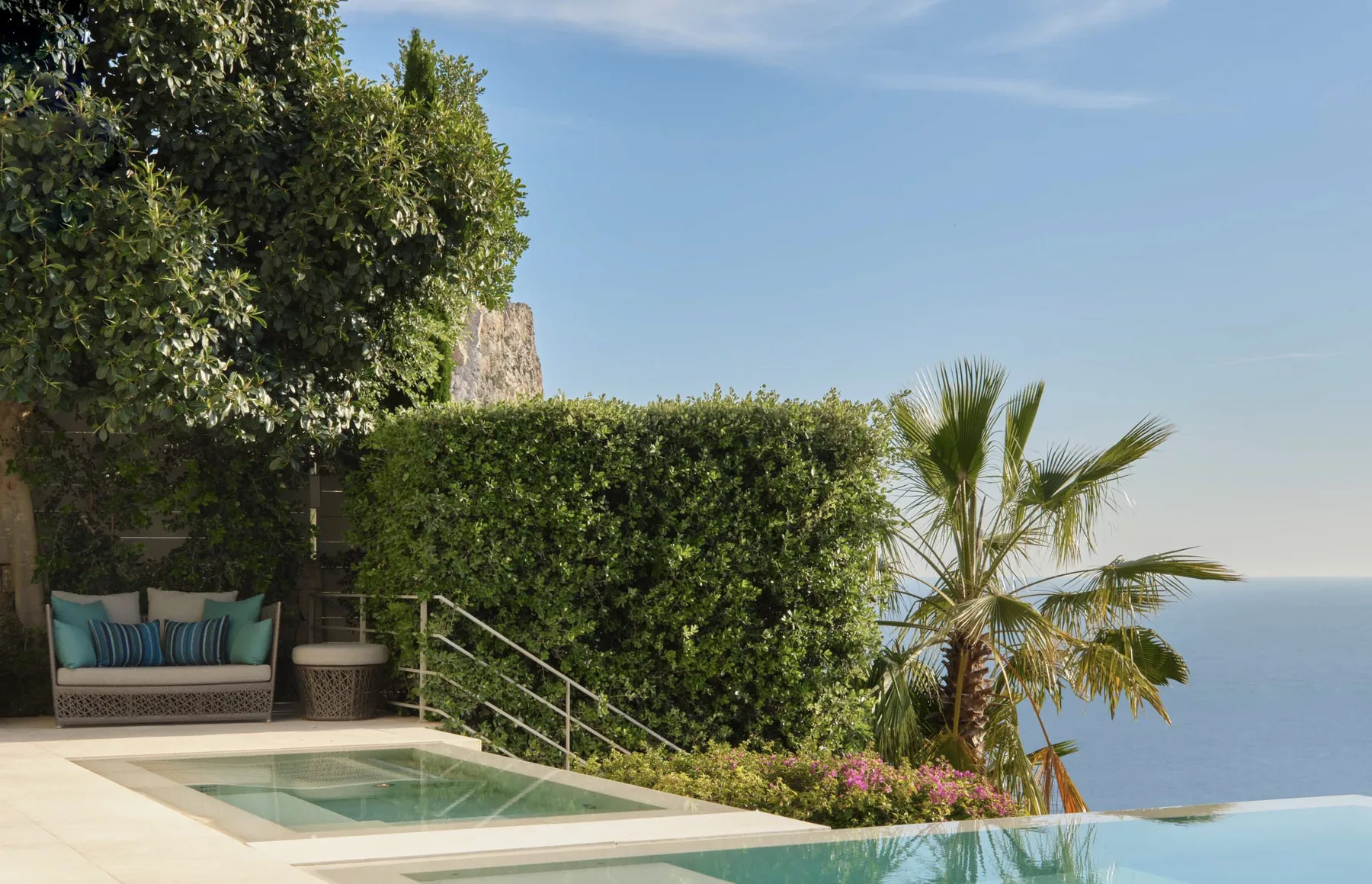 An outdoor pool beside a hedge, palm trees, and a seating area overlooks the sea.