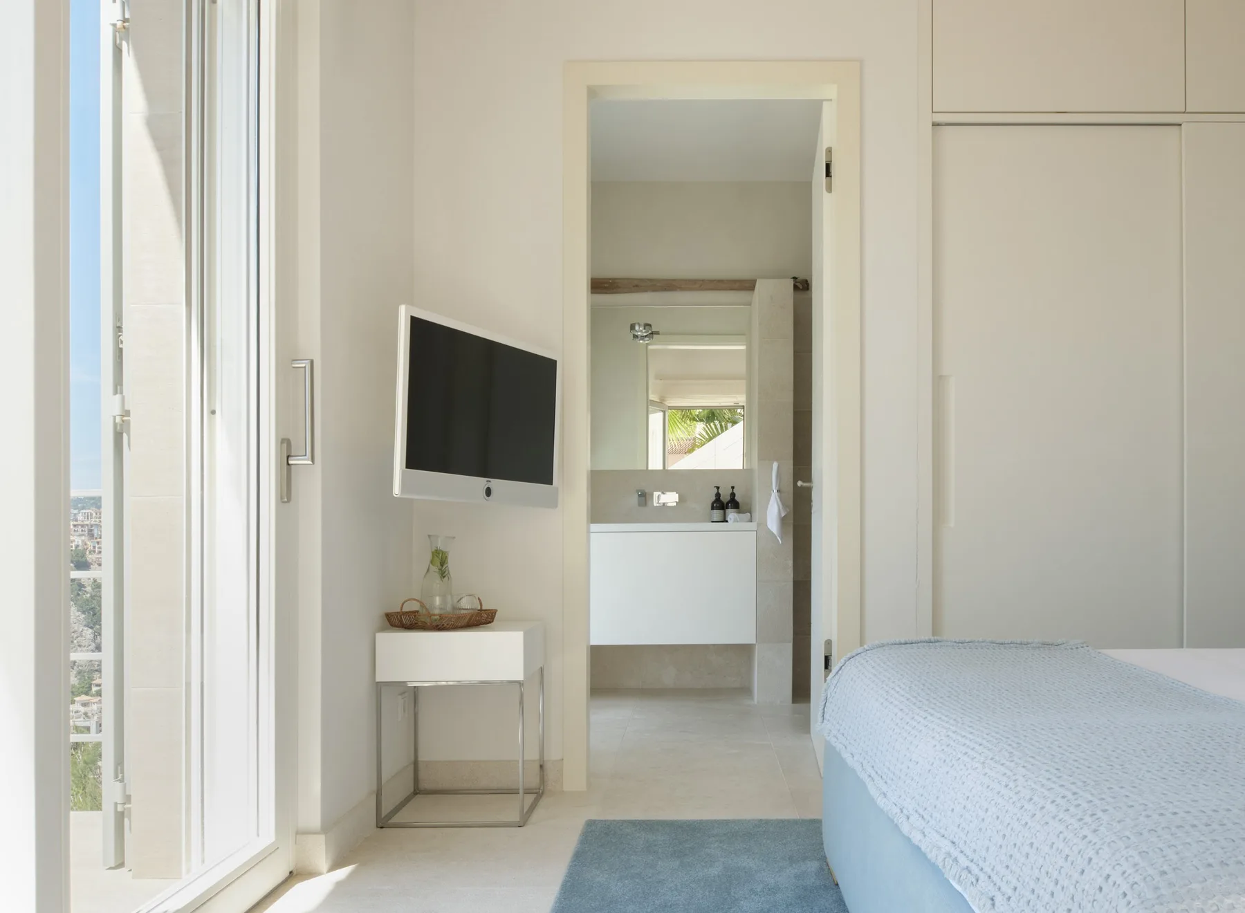 A bright bedroom with a bed, wall-mounted television, and an open doorway to the bathroom.