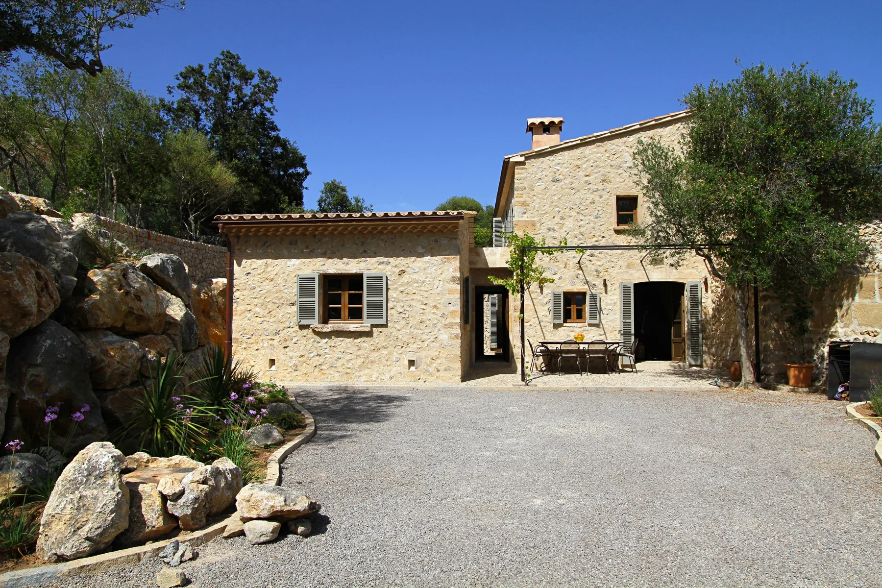 Una casa de piedra da a un amplio patio de grava, con árboles y rocas cerca.