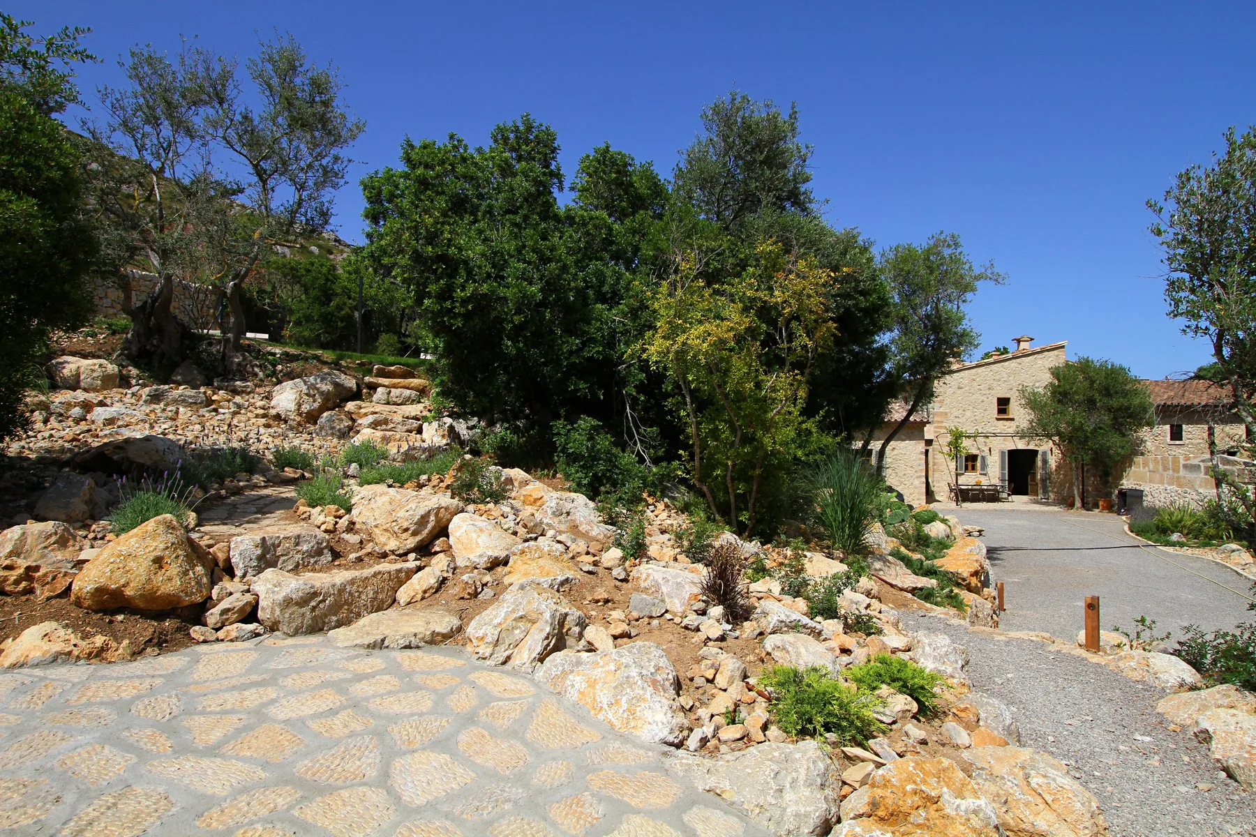 Un camino de piedra pasa junto a un jardín rocoso y árboles, hacia un pequeño edificio de piedra.
