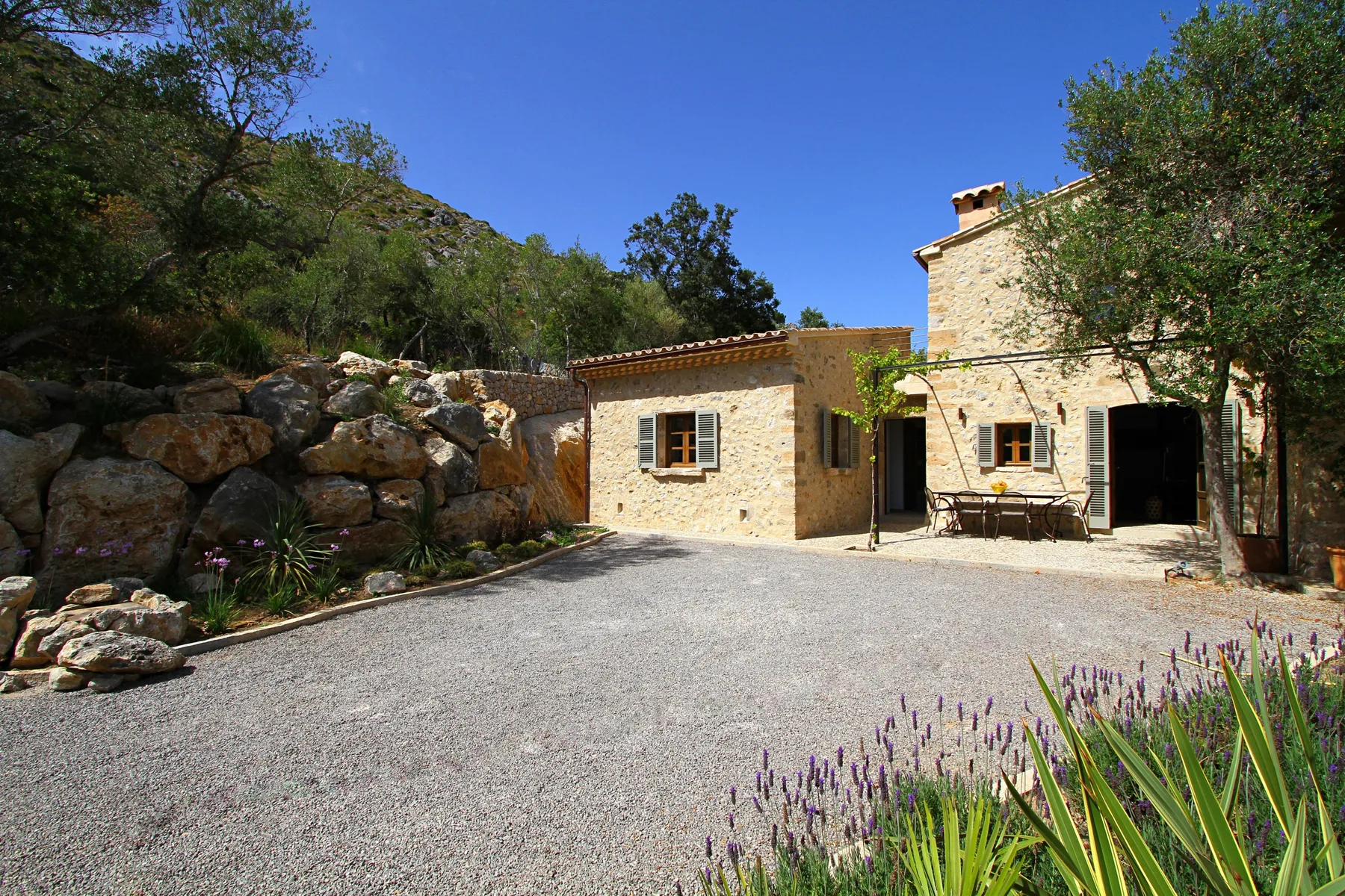 Una casa de piedra da a un patio de grava bordeado de rocas y plantas.
