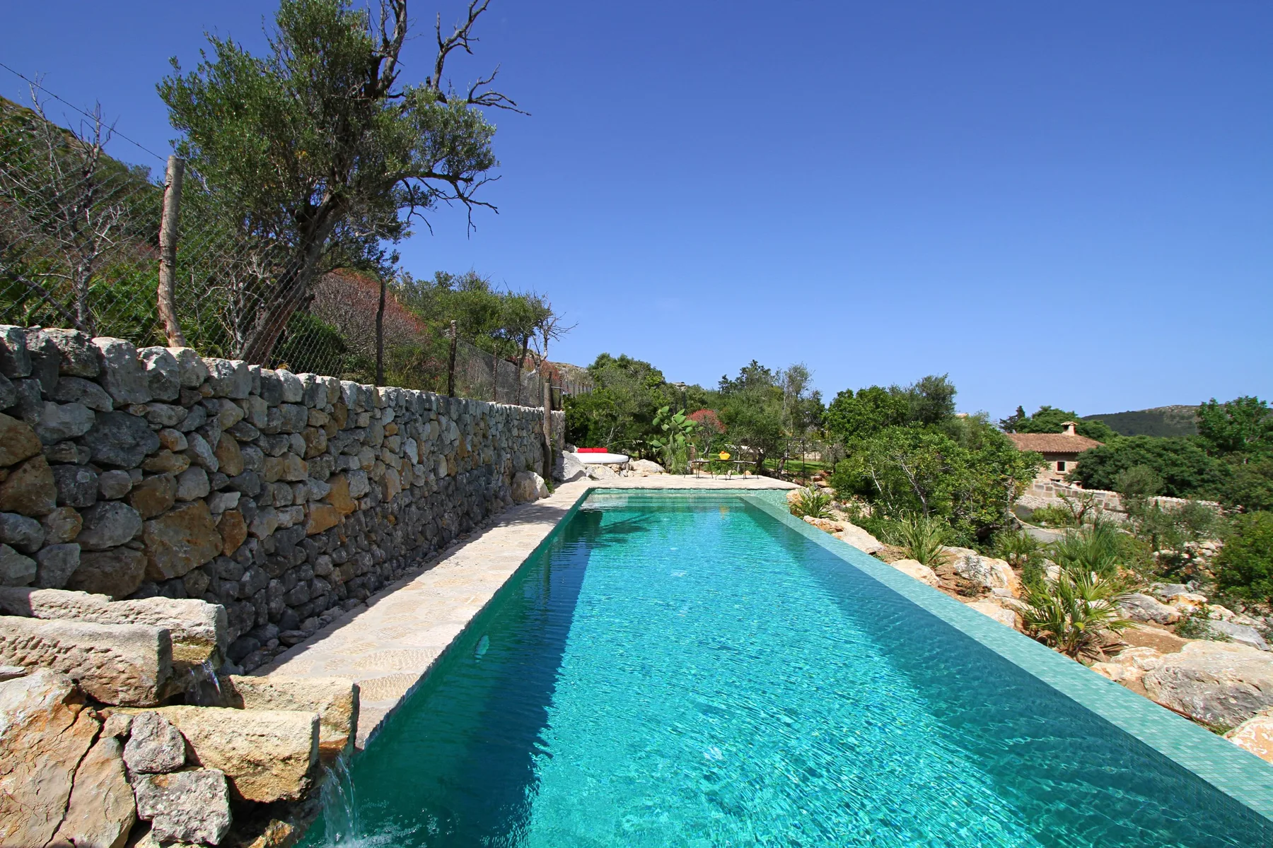 Una piscina turquesa está junto a un muro de piedra y un jardín.