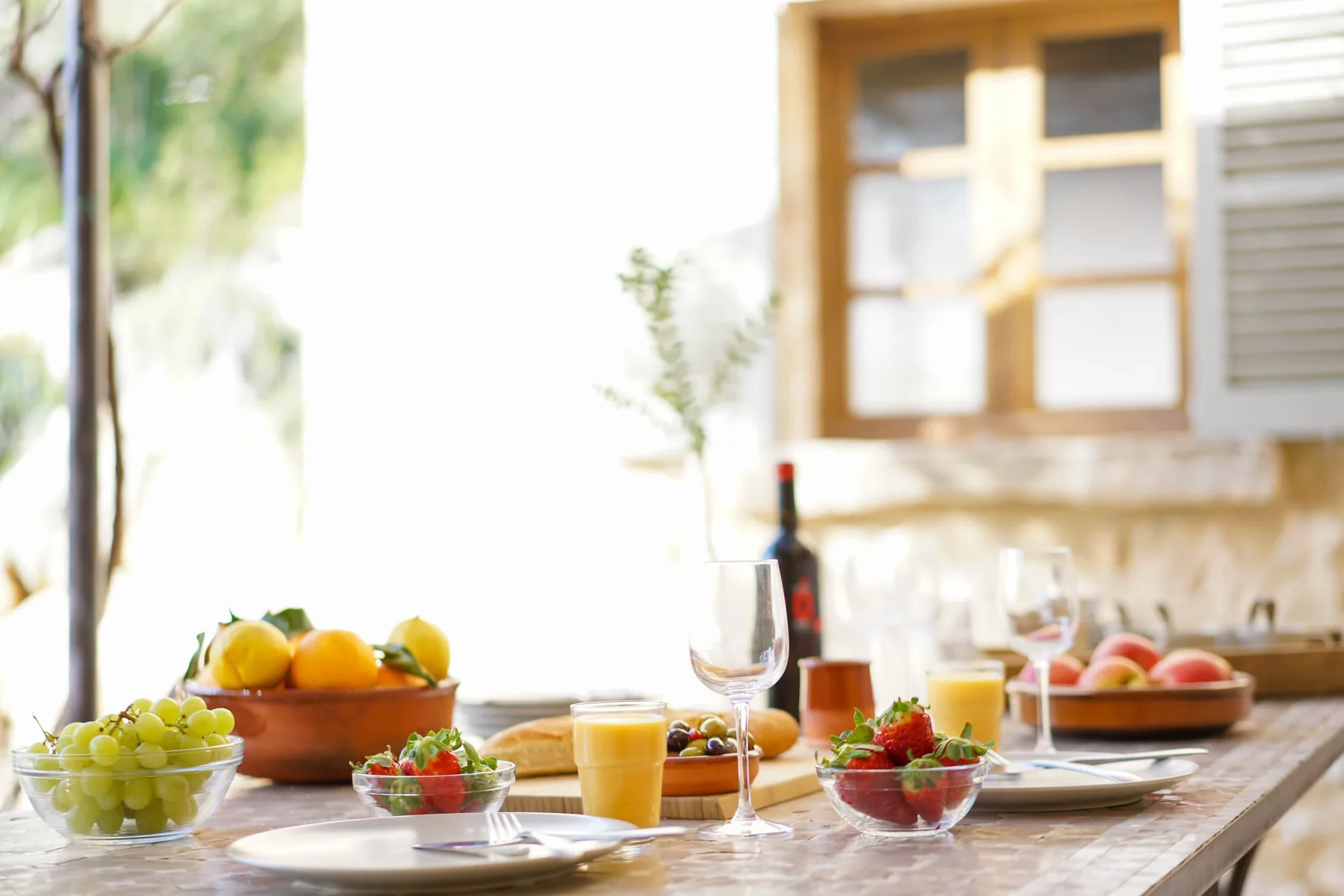 Una mesa de comedor luminosa está servida con fruta, jugo y copas de vino.