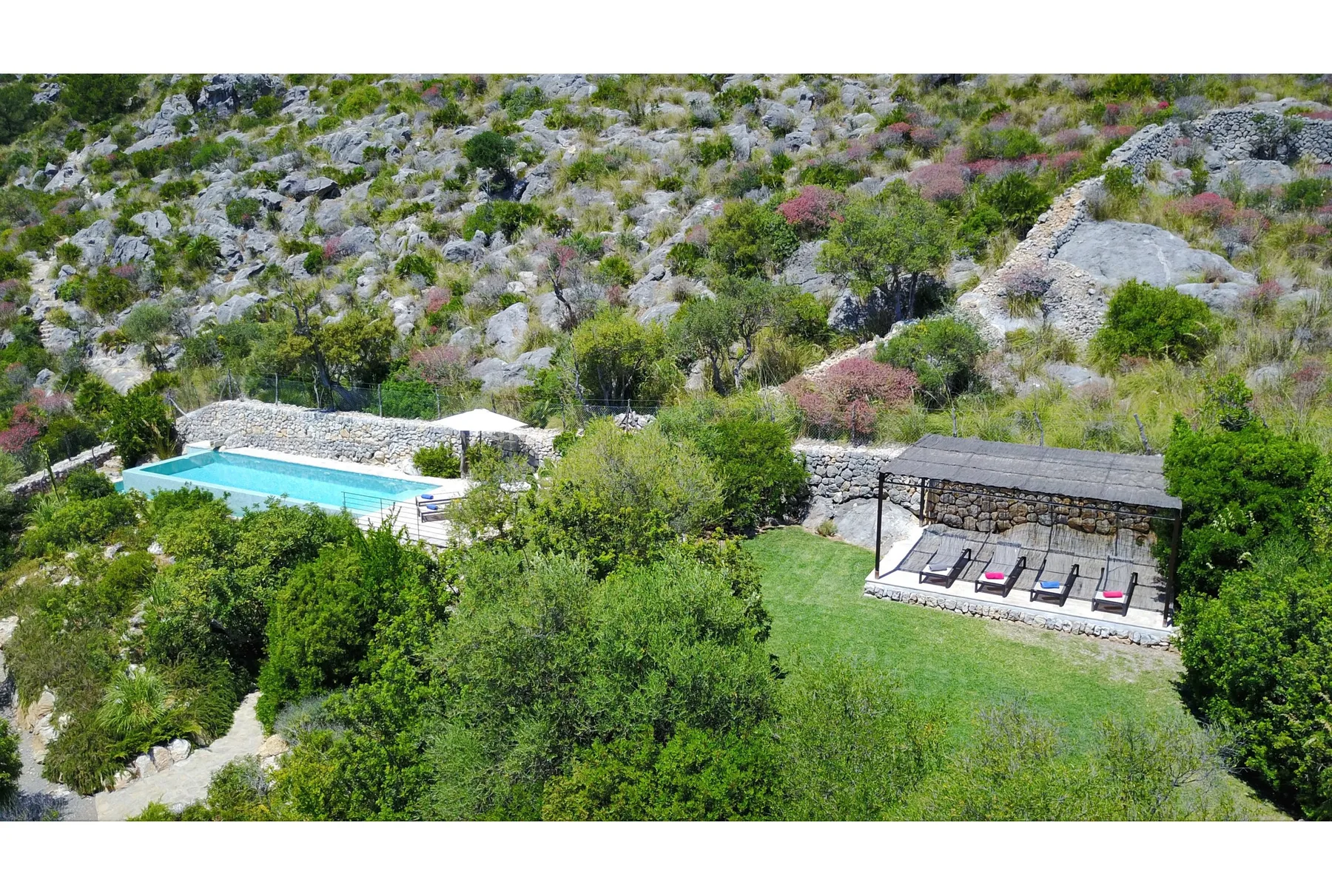 Una piscina al aire libre y una zona de estar sombreada están entre vegetación y colinas rocosas.