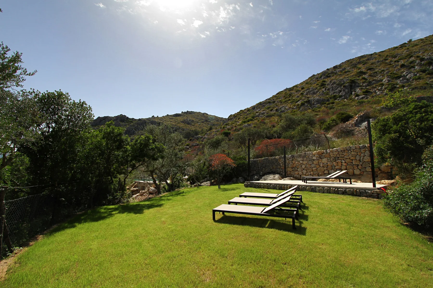 Un jardín soleado con césped, tumbonas y un muro bajo de piedra.