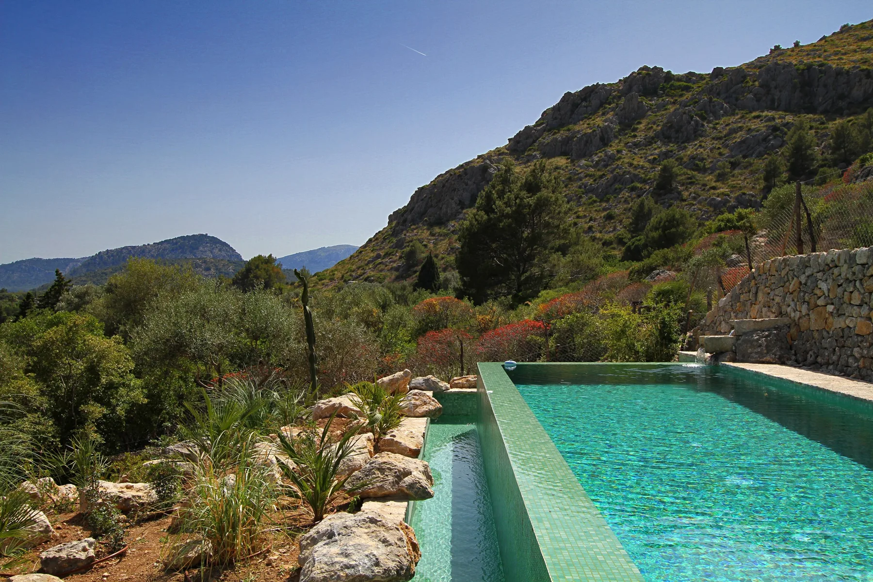 Una piscina exterior turquesa está junto a una ladera rocosa y vegetación verde.