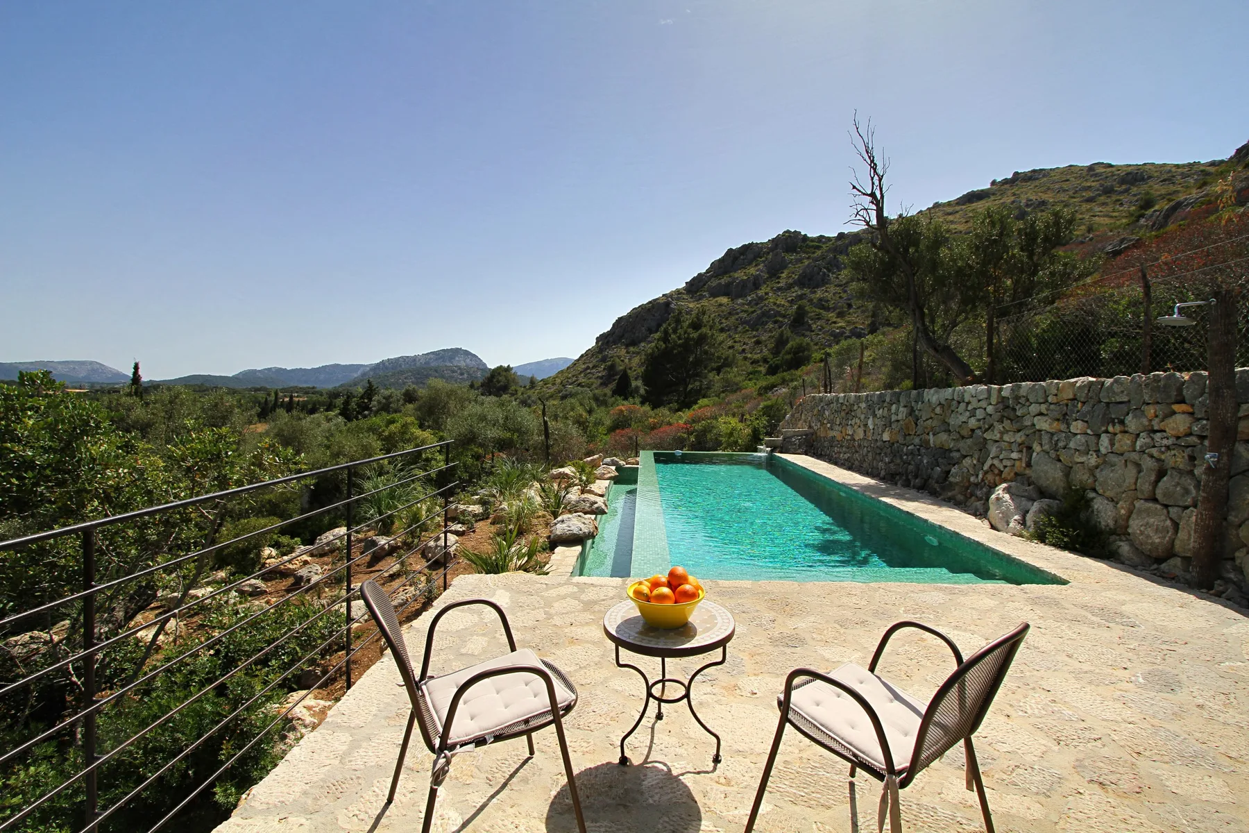 Una piscina junto a un muro de piedra, con asientos al aire libre frente a una ladera verde.