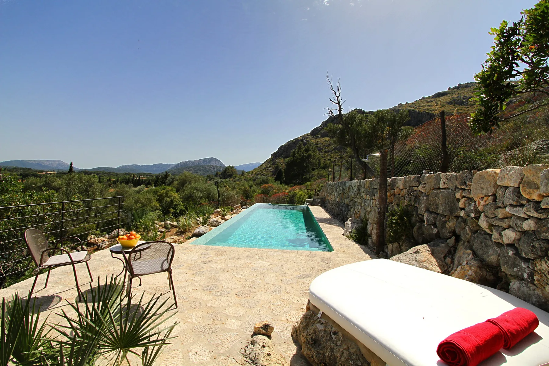 Una piscina al aire libre está junto a un muro de piedra, con asientos y colinas al fondo.